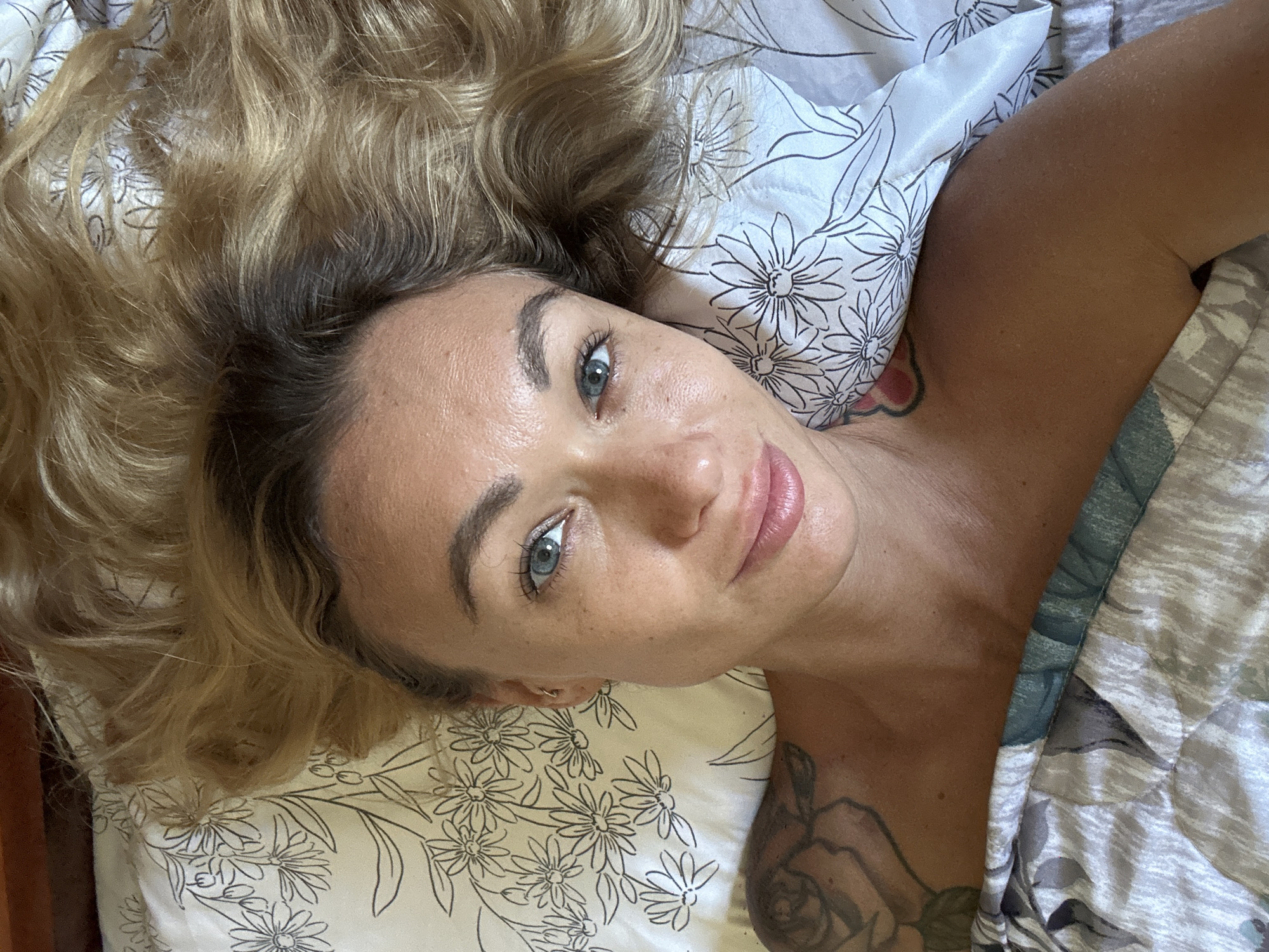 A woman with blonde, wavy hair lies on a bed with floral patterned sheets. She is looking up at the camera, with her head turned slightly to her left. Her eyes are blue and her lips are slightly parted. She is wearing a light-colored top, and a tattoo is visible on her left shoulder. The lighting suggests an indoor setting, possibly a bedroom. There is no indication of a specific location in Mũi Né, Vietnam.