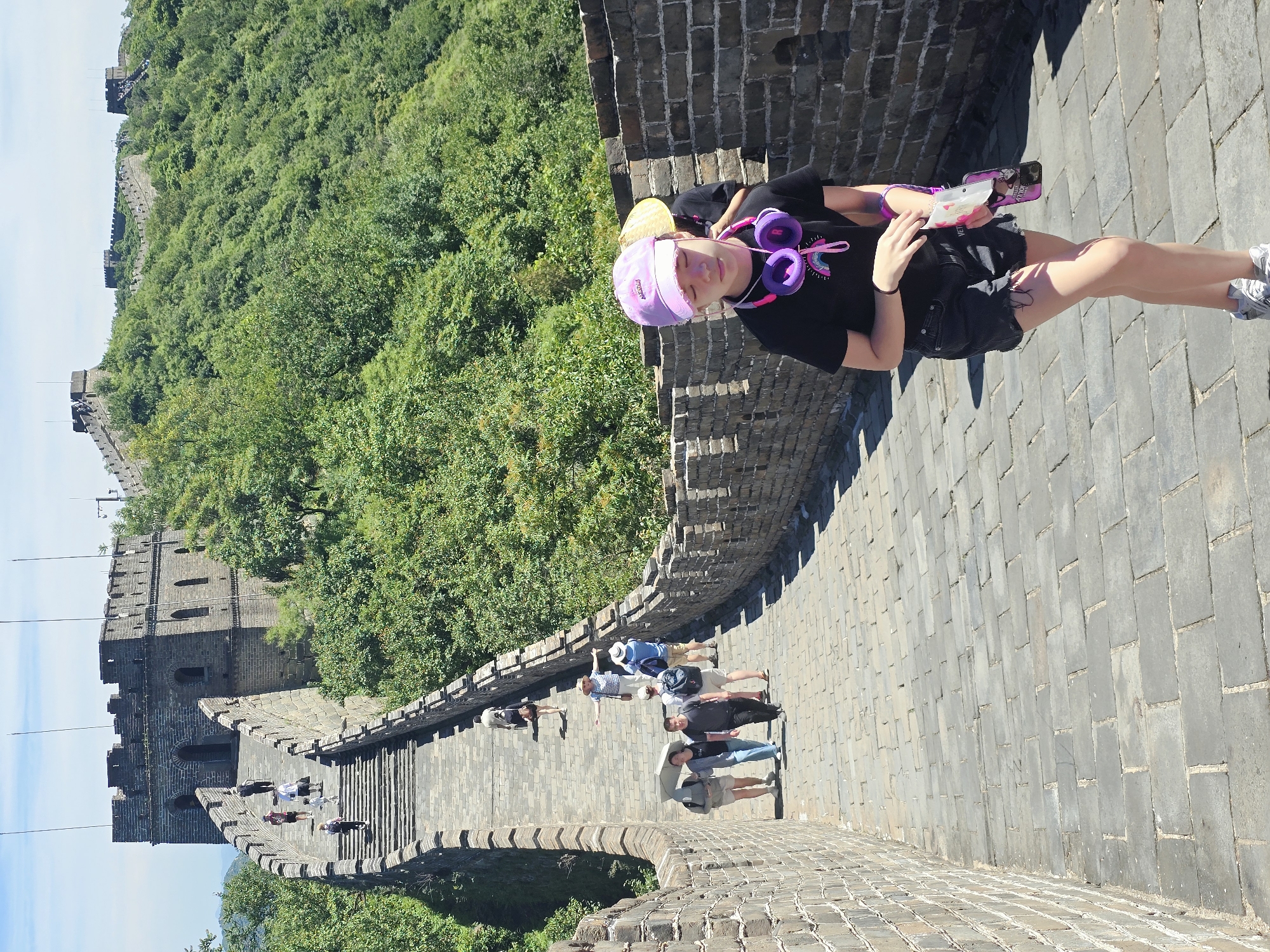 The image depicts a portion of the Great Wall of China, likely in the Huairou District of Beijing, China. The wall is constructed of stone and brick, with watchtowers visible in the background. The terrain is mountainous and covered in lush green trees.
In the foreground, a young girl with a purple baseball cap and headphones around her neck is walking down the wall. She is wearing a black t-shirt and dark denim shorts. She is holding a phone case with a character on it and a packet of tissues.
Further down the wall, a group of tourists are walking in both directions. Some are carrying umbrellas, and others are wearing hats. The wall appears to be a popular tourist attraction. The sky is clear and blue.
