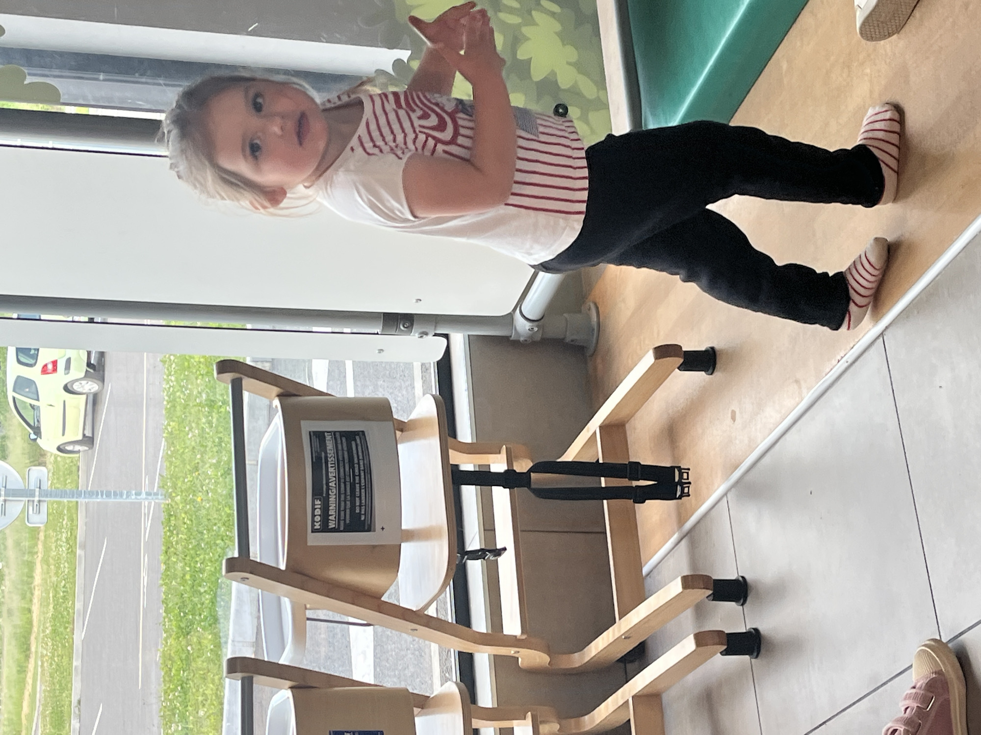 A young girl stands adjacent to a wooden high chair within an indoor setting. The high chair bears a warning label in both English and French, "WARNING/AVERTISSEMENT." The girl wears a striped shirt and black pants with striped socks. Outside the window is a parking area and a vehicle. Based on the "KODIF" branding on the high chair and warning label, the location is Andrézieux-Bouthéon, France.