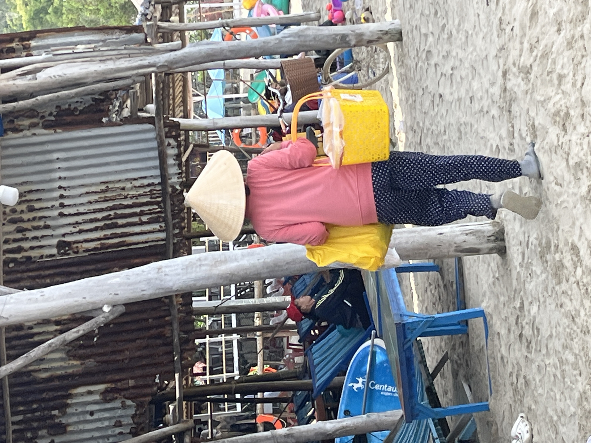 A person wearing a conical hat and a pink long-sleeved shirt is walking on a sandy beach in Nam Nha Trang Ward, Vietnam. They are carrying a yellow plastic basket with a white lid and a yellow bag over their left shoulder. The person is wearing dark pants with a white polka dot pattern and light-colored sandals. In the background, there are several blue lounge chairs, some with blue cushions, and a surfboard. A person in a red cap is sitting in one of the lounge chairs. Further back, there is a structure made of corrugated metal and wooden poles, with umbrellas and other beach equipment visible. A bicycle with a wicker basket is also present. The environment is a beach with sand, and some vegetation is visible in the distance.
