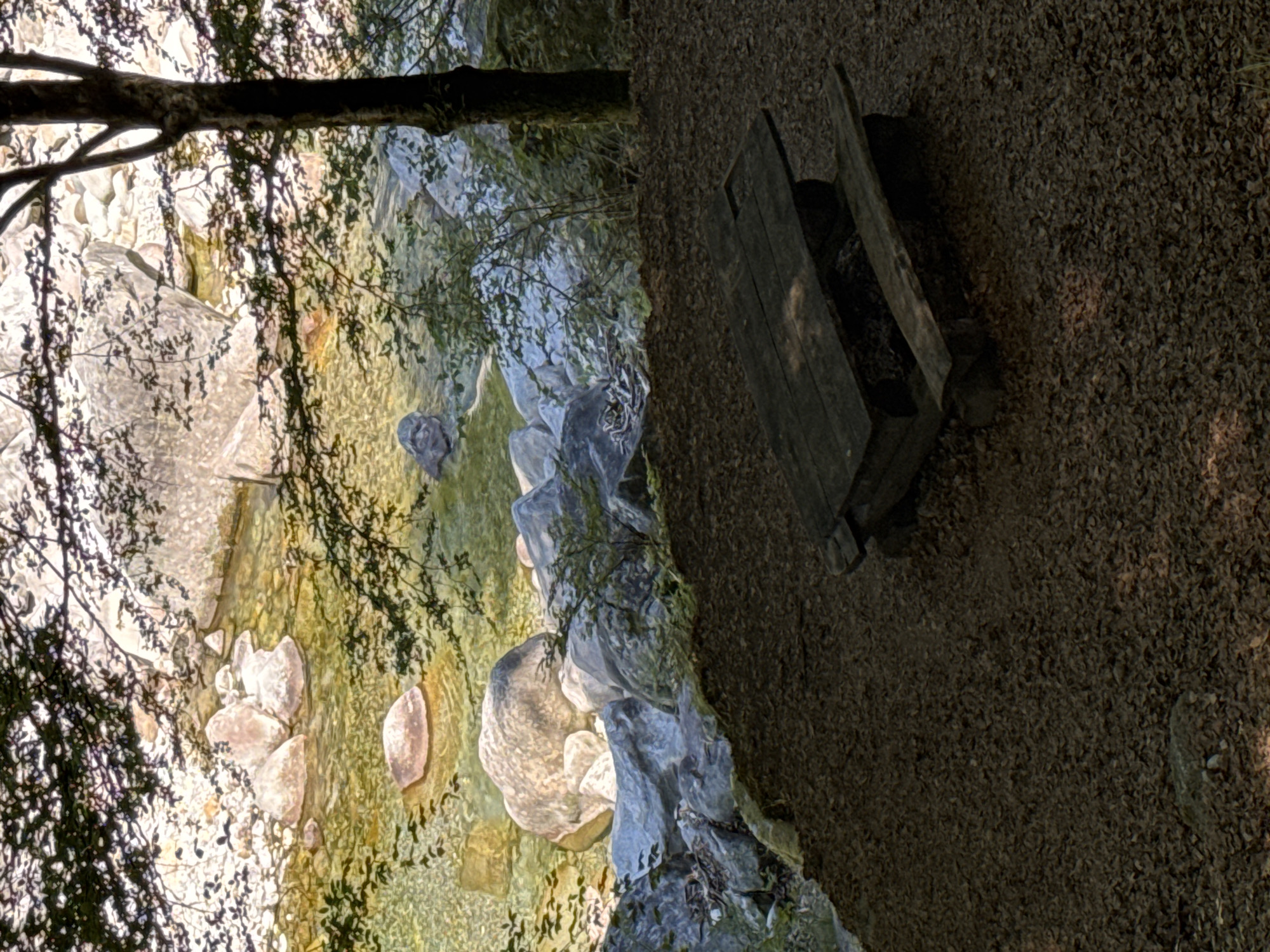 The image depicts a rustic wooden picnic table and benches situated on a ground covered with brown leaves, adjacent to a clear, shallow river flowing over smooth, grey and tan rocks. Overhead, the dappled sunlight filters through the branches of a tree, casting shadows on the scene. The river itself is characterized by its crystalline water, allowing the riverbed and submerged rocks to be visible. The background features larger boulders and lush vegetation, hinting at a natural, forested environment. The picnic table appears weathered, suggesting it has been exposed to the elements for some time. The overall atmosphere is tranquil and natural, typical of an outdoor recreational area in a mountainous or woodland setting. The location is identified as being in the city of Verzasca, Switzerland, a region known for its scenic beauty and clear rivers.