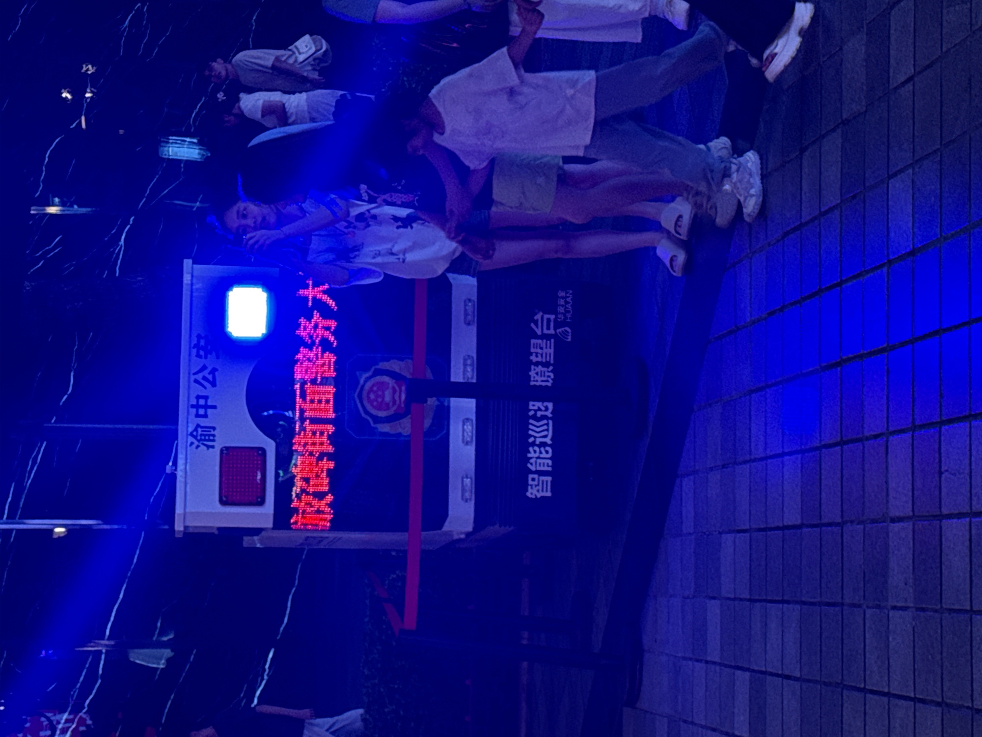 In Yuzhong District, China, a police vehicle with blue flashing lights is parked on a tiled sidewalk at night. The vehicle has Chinese characters on it, including "渝中公安" (Yuzhong Public Security) and "欺峰街面警务" (Qifeng Street Police Service). A digital display on the vehicle shows red Chinese characters. Several people are standing in front of the vehicle, some looking at the display or the camera. The environment is lit by blue lights, casting shadows on the ground and surrounding surfaces, which appear to be a dark, reflective wall or structure.