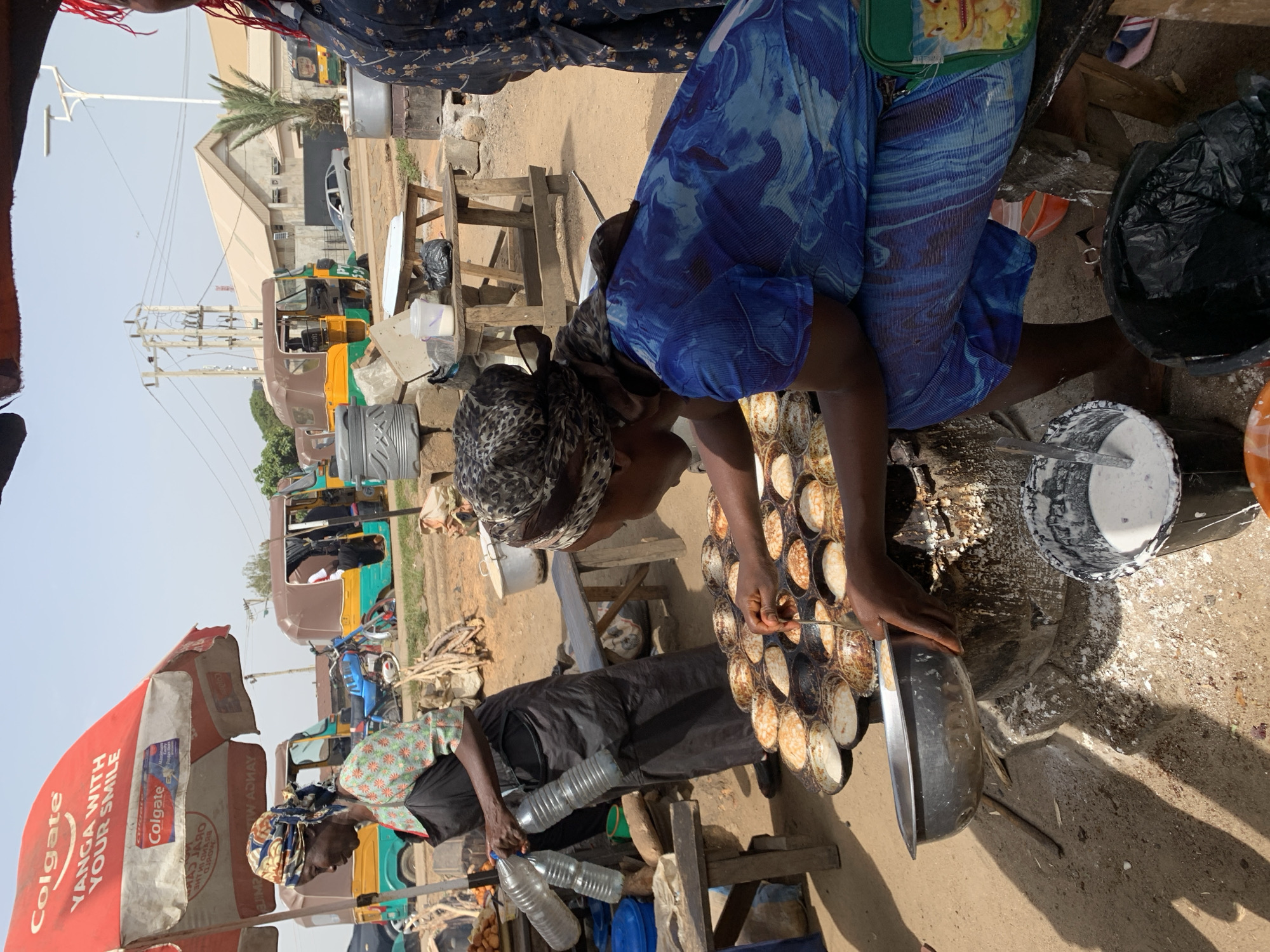Two women are preparing food on the street in Rafin Par, Nigeria. One woman is wearing a blue patterned dress and leopard print headscarf, bending over a pan with multiple round indentations, filling each with a batter-like substance using a small utensil. Beside her is a bucket containing the batter. The second woman is seen under a Colgate umbrella, wearing a green patterned top and black pants. She is pouring water from plastic bottles. In the background, there are parked auto-rickshaws and various structures. The ground is bare, with some scattered debris.