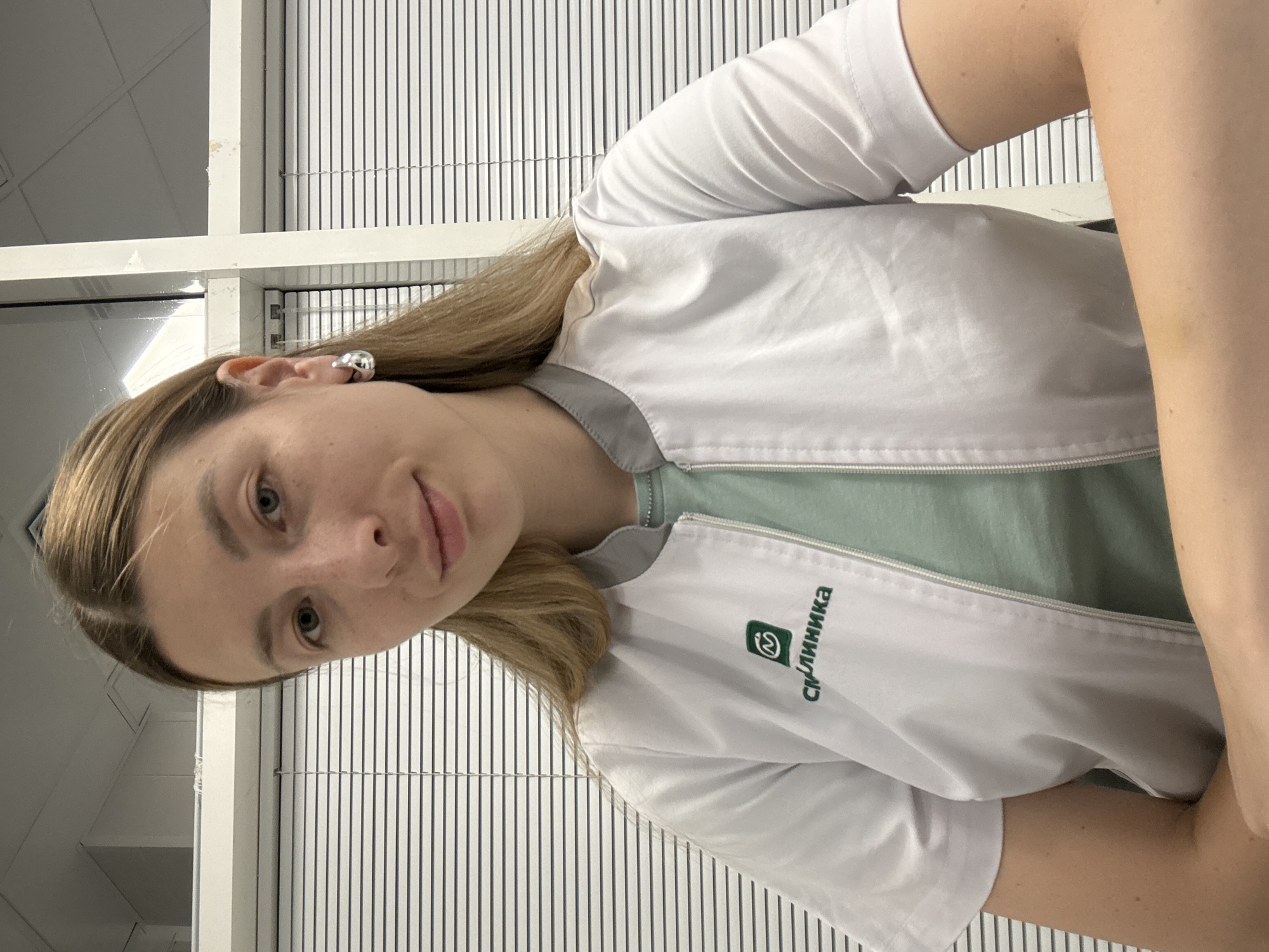 A young woman with long blonde hair is looking at the camera with a slight smile. She is wearing a white lab coat over a light green shirt. The lab coat has a green logo with Cyrillic text that reads "СМ. КЛИНИКА". She is also wearing a silver hoop earring in her left ear. The background appears to be an office environment with white blinds covering windows. The image was taken in Moscow, Russia.