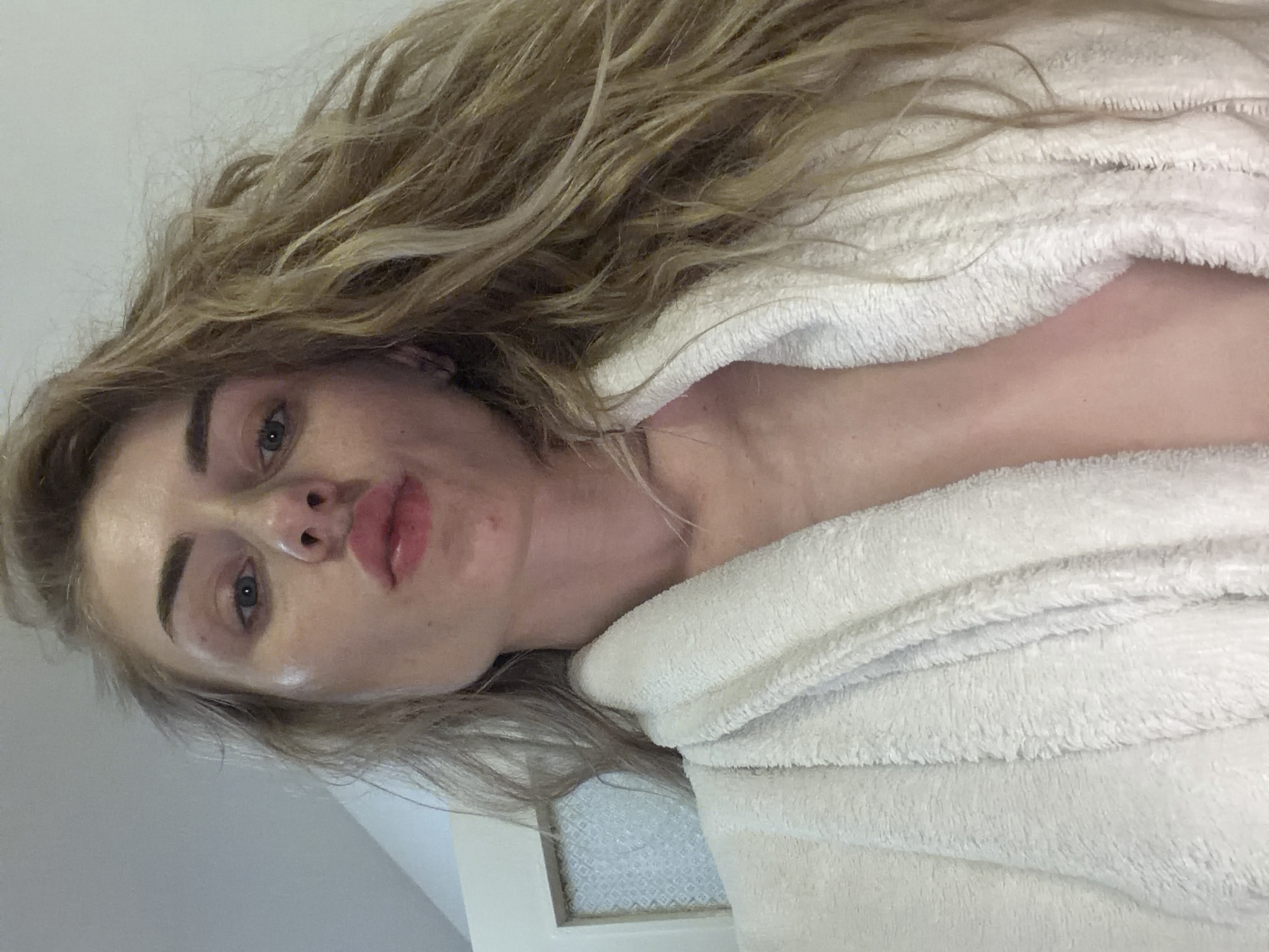 An overhead, chest-up medium shot captures a young woman in a white terrycloth bathrobe. She is looking directly at the camera with her lips puckered. Her blonde, wavy hair is styled casually and falls over her shoulders. The background is out of focus, with a hint of a door on the left side of the frame.