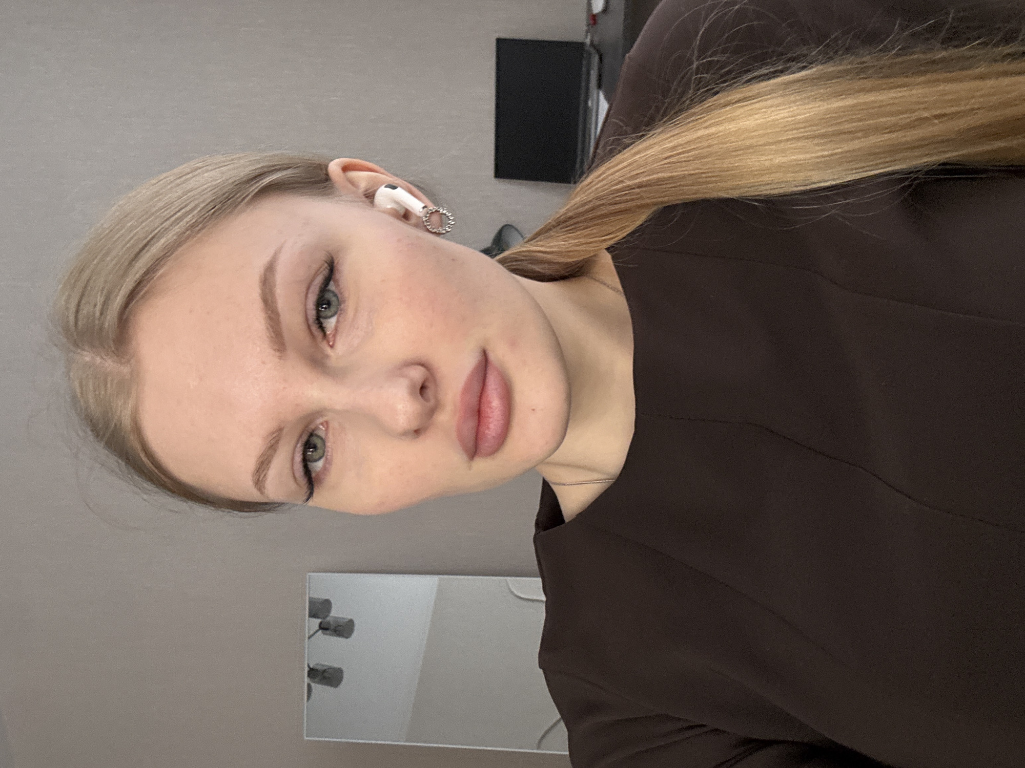 A close-up, slightly angled portrait depicts a young woman with light blonde hair, fair skin, and light-colored eyes. She is wearing a dark brown or black top. In her left ear is a white earbud with a decorative, possibly rhinestone, ring around the base. The background is a muted gray. A dark, rectangular object is partially visible behind her, and the top of a light-colored rectangular object is seen in the lower portion of the image, potentially a mirror and the corner of a TV. It is not possible to definitively locate the image as being from Perm, Russia.