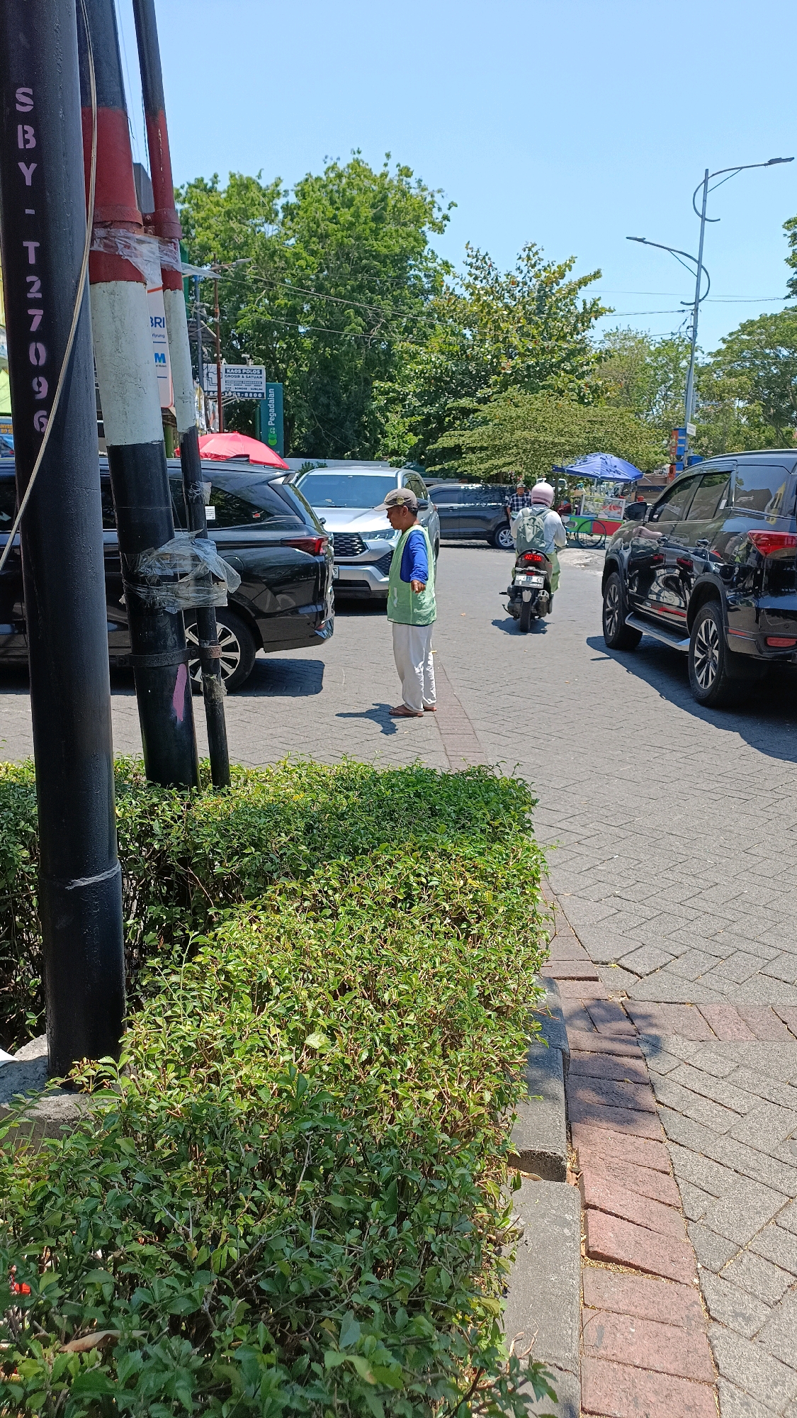 This image captures a street scene in Surabaya, Indonesia, likely near a commercial area given the presence of cars and shops. In the foreground, a person wearing a light green vest over a blue shirt and light-colored pants stands on a paved sidewalk next to a low, dense hedge. The person is looking to their left, with their right arm extended slightly. Behind this individual, several vehicles are visible, including a black SUV and a silver car. Further down the street, a person is riding a motorcycle, also heading away from the viewer. The background features lush green trees and a clear blue sky. Traffic appears moderate, and the overall impression is of a sunny day in an urban environment. The presence of a pole with signage, partially obscured by a black post, suggests this is a public street.