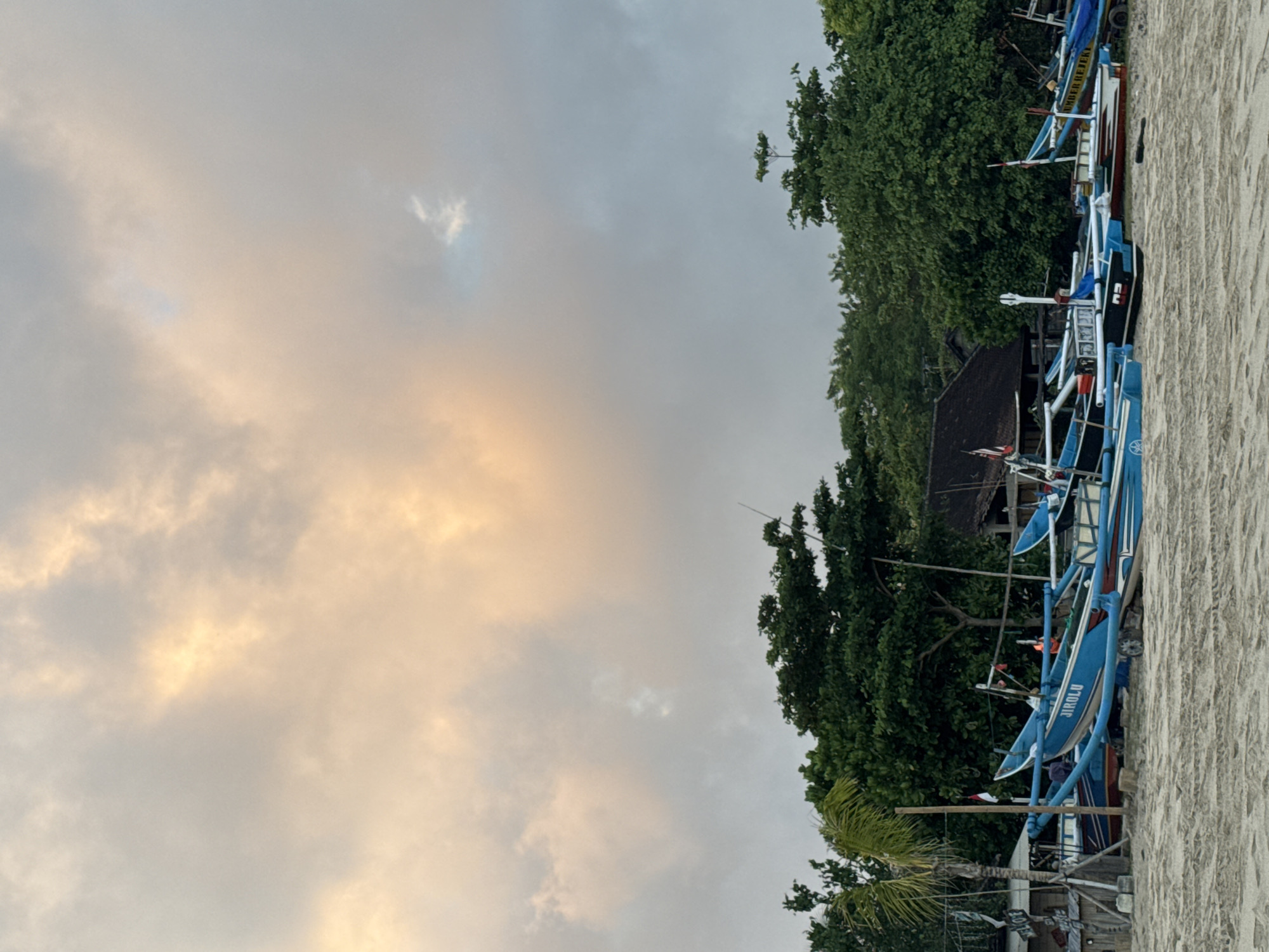 This is a wide shot of a beach in Kuta, Indonesia, with a line of brightly colored fishing boats pulled up onto the sand. The sky above is a mix of soft, warm clouds in shades of peach and yellow, contrasted with cooler gray tones, suggesting either sunrise or sunset. Lush green trees form a dense backdrop behind the boats, with a hint of a building structure peeking through the foliage. The sand in the foreground shows textured patterns, likely from the receding tide or footprints. The boats are the primary subjects, with visible names and markings on some of them, such as "JIROLU" on one blue hull. There are no people visible in the frame. The overall scene conveys a peaceful, tropical coastal atmosphere.