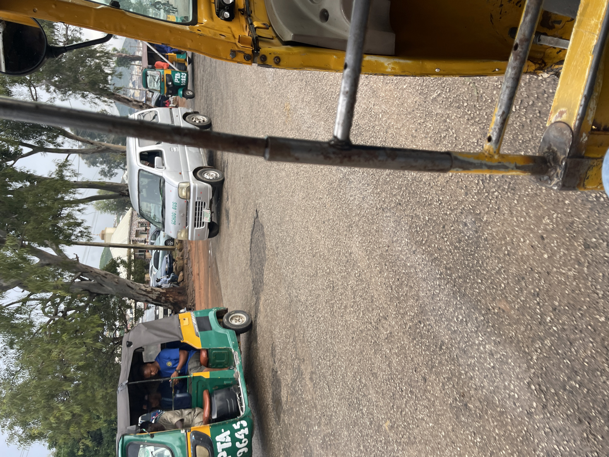 This media file captures a scene on a paved road, likely in Jos, Nigeria, as indicated by the location context. The dominant elements are vehicles, including several auto-rickshaws (also known as Keke Napep) and a white van.
In the foreground, partially obscuring the view of the road, is the interior of a yellow auto-rickshaw. We see metal bars from its frame, suggesting the perspective is from within or beside one of these vehicles.
Further down the road, a green and yellow auto-rickshaw is prominently visible. A man wearing a blue shirt with a circular emblem is seated in the driver's position, appearing to be in control of the vehicle. Another person is seated behind him, and partially visible in the front passenger seat is another individual. Visible text on this auto-rickshaw includes "PTA-9645".
To the left of this auto-rickshaw, a white van is parked. Text on the side of the van reads "SCHOOL BUS". Behind the van, other vehicles and buildings are visible, suggesting an urban or semi-urban environment. There are also several trees, with one large, prominent tree with sparse foliage dominating the left side of the frame.
The road surface is asphalt, with some visible cracks and imperfections. The lighting suggests it is daytime, and the weather appears to be overcast or hazy, as there are no strong shadows. The overall atmosphere is that of a typical day with vehicles on the road, possibly in traffic or preparing to move. No specific emotions are conveyed by the individuals, as they are either partially visible or not the primary focus.