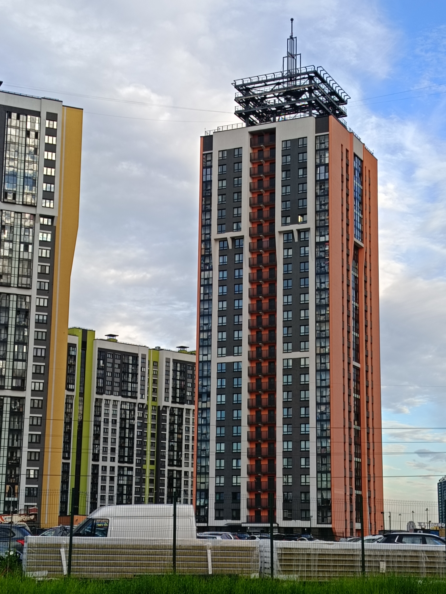 This is a wide-angle, eye-level, outdoor, daylight photograph of a cluster of modern high-rise apartment buildings in Saint Petersburg, Russia. The buildings feature a mix of architectural styles and colors, with large windows, balconies, and a prominent antenna structure atop one of the tallest buildings. In the foreground, a white van and several parked cars are visible behind a chain-link fence, with stacks of building materials in front of the fence. The sky is cloudy with patches of blue.