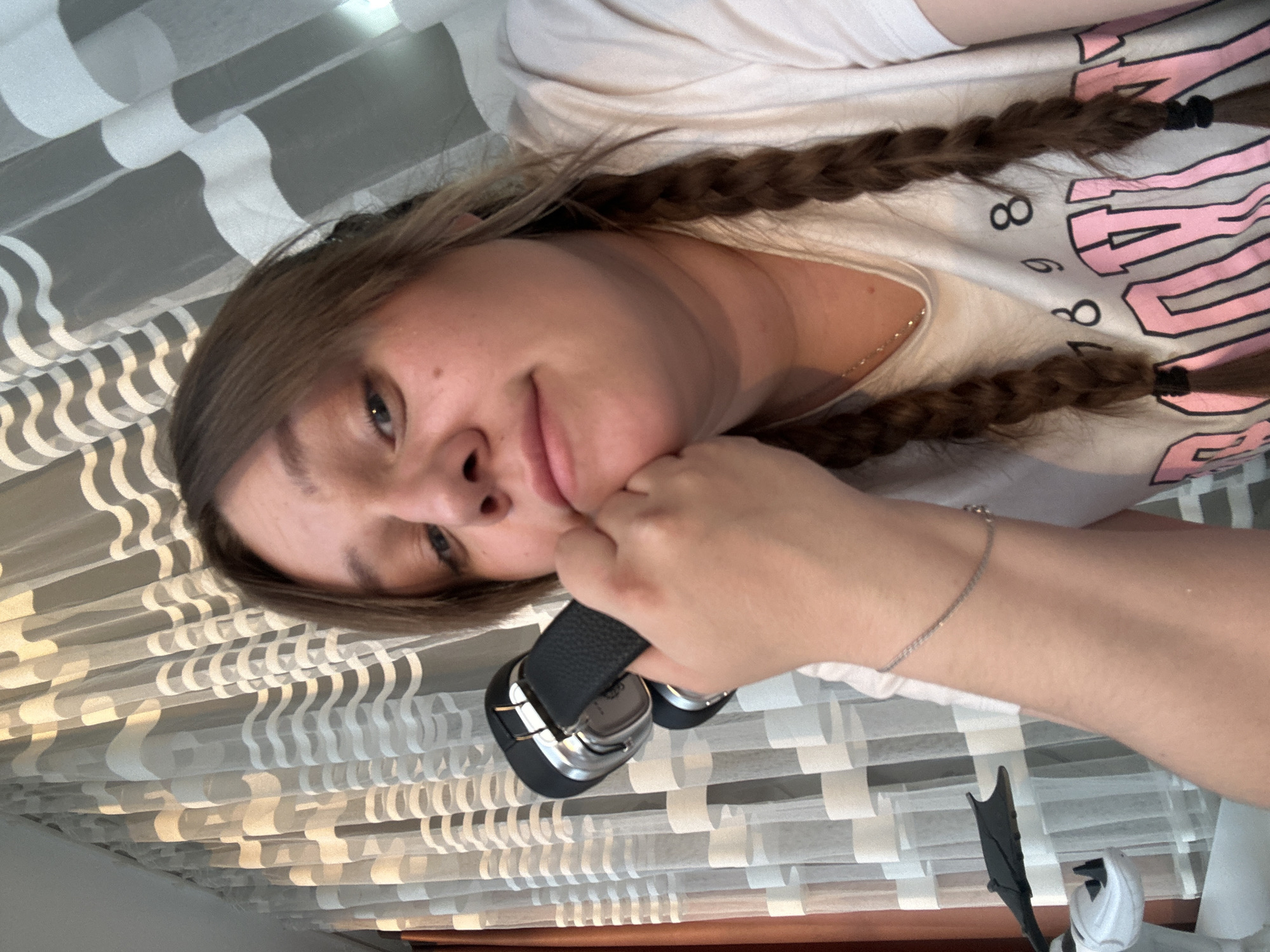 A woman with blonde hair styled in two braids is holding black and silver headphones to her right cheek. She is wearing a light-colored t-shirt with pink lettering and a silver chain necklace. She is also wearing a silver bracelet on her right wrist. Behind her is a textured, white and gray striped curtain. The image includes a partial view of a white and black figure at the bottom right. Based on the available information and scene context, I cannot pinpoint the exact location in Chaikovsky, Russia.