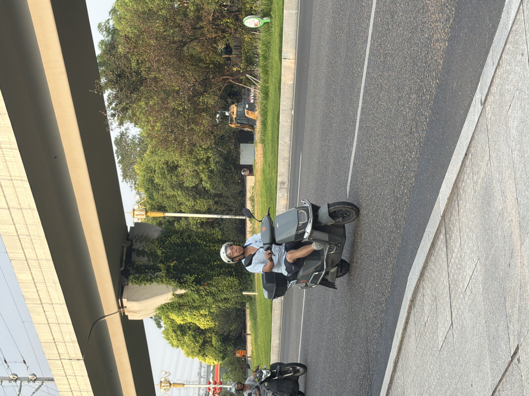 A medium shot shows two individuals riding motor scooters on a paved road under an elevated highway. The scooter in the foreground is ridden by a person wearing a blue shirt and shorts, a white helmet, and a face shield. This scooter is a dark grey color with a black storage box mounted on the rear. The second scooter, in the background and to the left, is being ridden by a person wearing a light grey top and a helmet. This scooter is black and has a windshield. The road surface is asphalt, with white lane markings visible. To the right of the road, a grassy median separates the road from more greenery and trees. An excavator is partially visible in the background among the trees. The elevated highway structure is made of concrete, with concrete support pillars partially covered in green vines. The sky is clear and bright. The scene appears to be in a semi-urban or suburban area of Jiangjin, China.