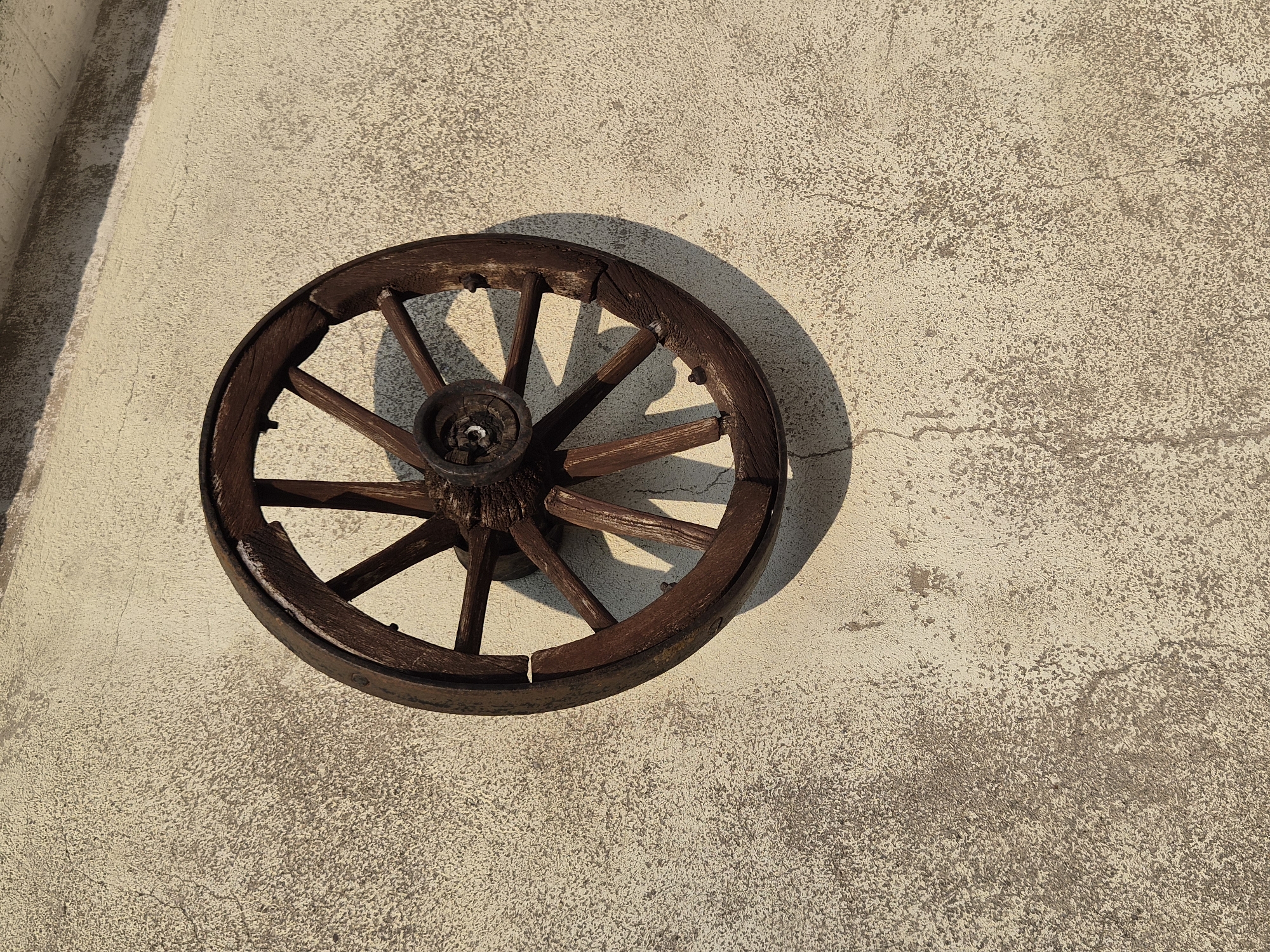 An antique wooden wagon wheel, weathered and dark brown, is affixed to a textured, light-colored stucco wall. The wall shows signs of age and possibly water damage, with a rough, mottled surface and some faint cracks. The wheel, with its spokes and rim, casts a distinct shadow on the wall, indicating a strong light source, likely the sun, from the left. The scene is devoid of any people or animals, and there are no discernible elements that would suggest a specific location within the city of Eisenerz, Austria, beyond being an exterior wall. The focus is solely on the static composition of the wheel against the textured surface.