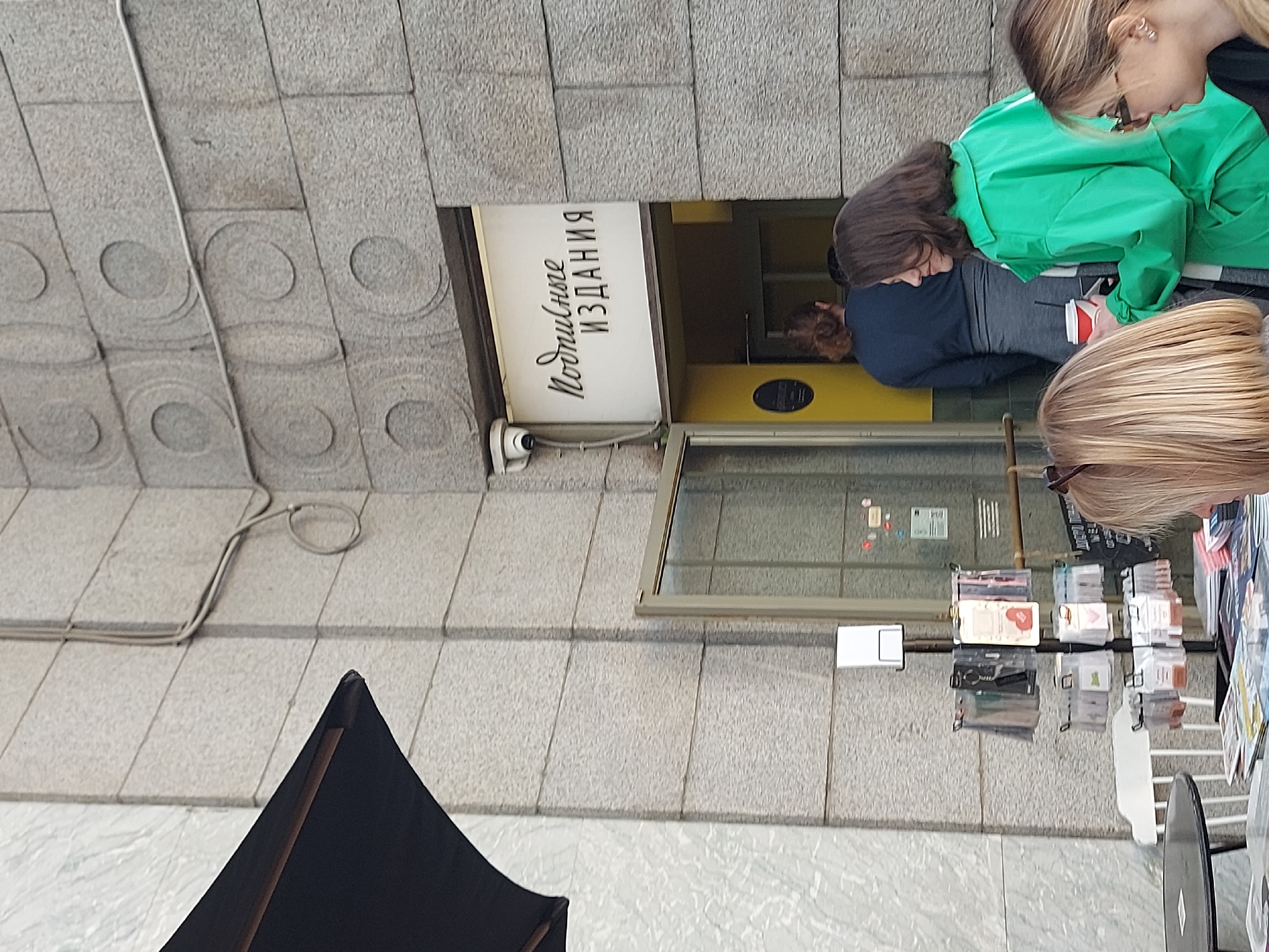 The image depicts a street scene in Saint Petersburg, Russia. The focus is on the entrance to a business, presumably a publishing house or bookstore, as indicated by the sign "Подписные издания" (Subscription Editions).
Three people are present, two women are looking towards a rack that is full of small books and or cards. Another person is inside the business, their face close to the door.
The environment features grey stone architecture, a common characteristic of Saint Petersburg. The area appears to be a pedestrian zone, with a portion of a black tent-like structure visible in the lower left corner.