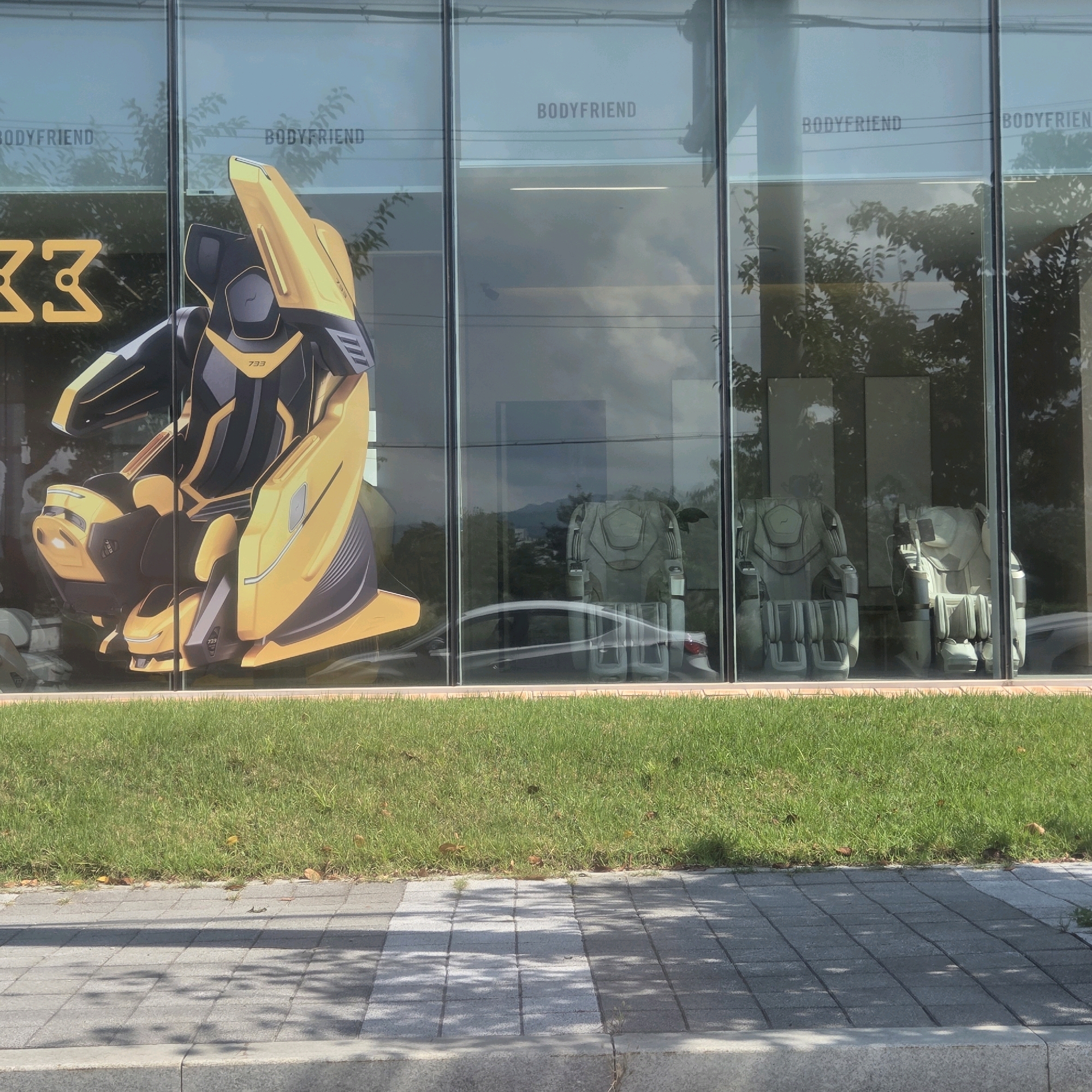 The image depicts a storefront with large glass windows, possibly in Gangneung-si, South Korea. A large graphic of a yellow and black futuristic seat is displayed on the left side of the window, with the number "33" visible. The words "BODYFRIEND" are printed repeatedly on the glass above the graphic and on subsequent panels. Inside the store, several massage chairs, light in color, are visible through the windows. The chairs appear to be arranged in a showroom setting. In front of the store, a strip of green grass separates the building from a paved walkway with a cobblestone-like pattern. Shadows from trees are cast on the walkway. The overall scene suggests a commercial establishment, likely a showroom for the brand "BODYFRIEND," specializing in massage chairs.
