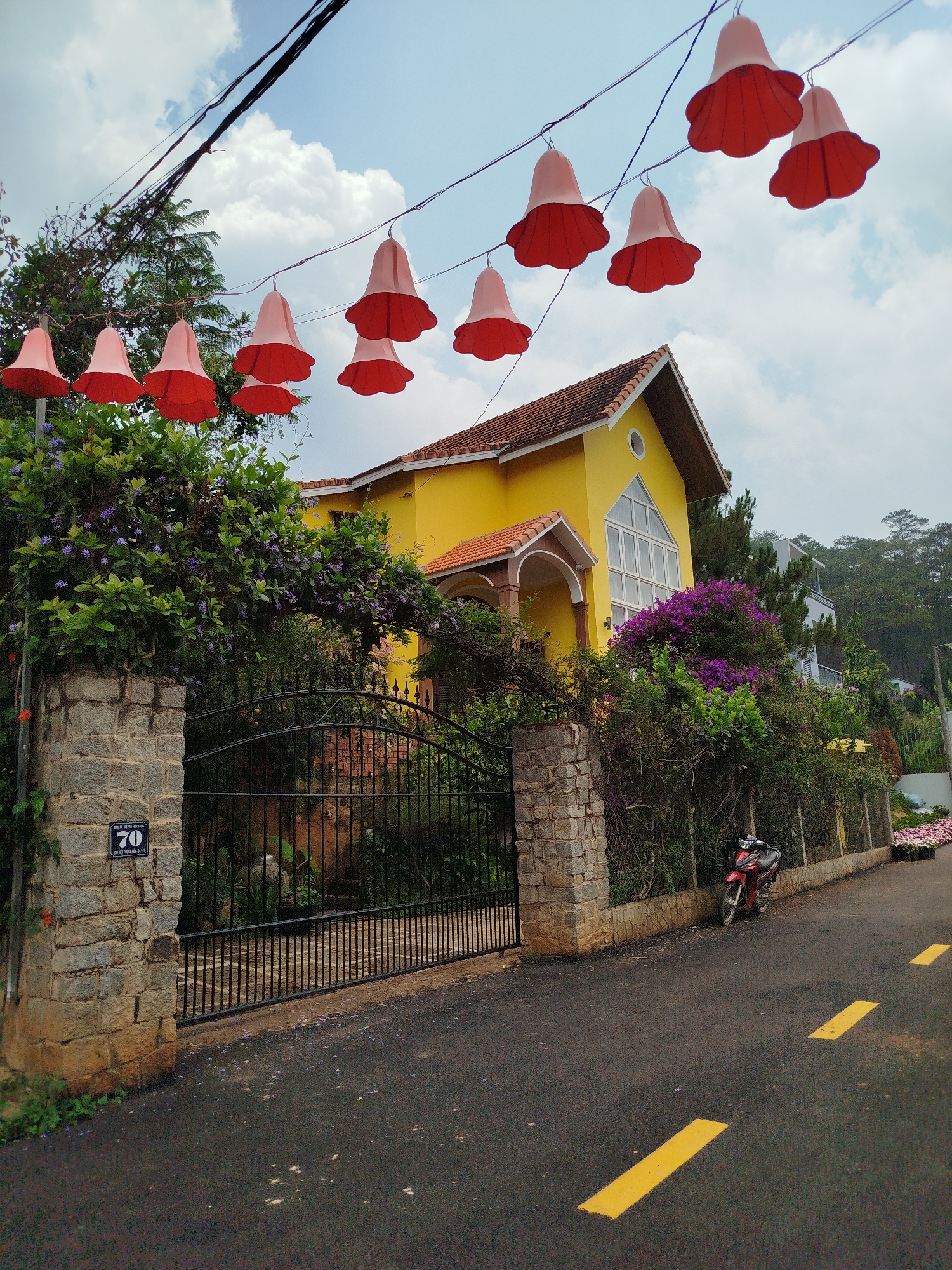 A yellow, two-story house stands behind a black wrought-iron gate and stone wall at 70 Truong Cong Dinh, Hiep Thanh, Vietnam. The house has a red-tiled roof, a large arched window, and a covered entryway. Lush greenery with purple flowers surrounds the property. A red motorbike is parked beside the fence. Above the road, a string of red bell-shaped decorations hangs, and yellow stripes mark the asphalt street. The sky is blue with scattered clouds.