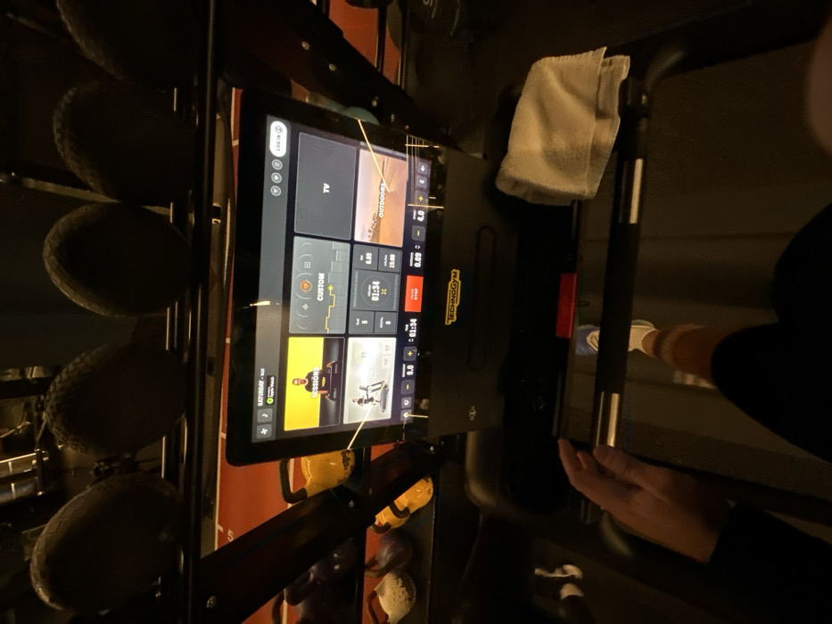 The image captures an indoor gymnasium scene from a first-person perspective, focusing on an individual utilizing a Technogym treadmill in London, United Kingdom. The prominent digital console of the treadmill displays a user interface, indicating "SATURDAY - 16:25" with an active Apple Watch connection. The screen presents various exercise options including "SESSIONS" (showing a male instructor), "CUSTOM," "TV," and "OUTDOORS," along with real-time workout metrics: a timer at "01:34," distance covered at "0.09" km, 0.0 elevation, and a speed of "4.0." A red "STOP" button is visible directly below the screen. The user's right hand rests on the left handle of the treadmill, while their lower right leg and a white sneaker are visible on the moving treadmill belt, indicating active exercise. A folded white towel is placed on the right side of the treadmill console. In the dimly lit background, a rack system holds several dark, spherical objects, consistent with medicine balls, above a shelf displaying multiple kettlebells in various colors (yellow, white, dark). An orange line marking is discernible on a surface in the background.