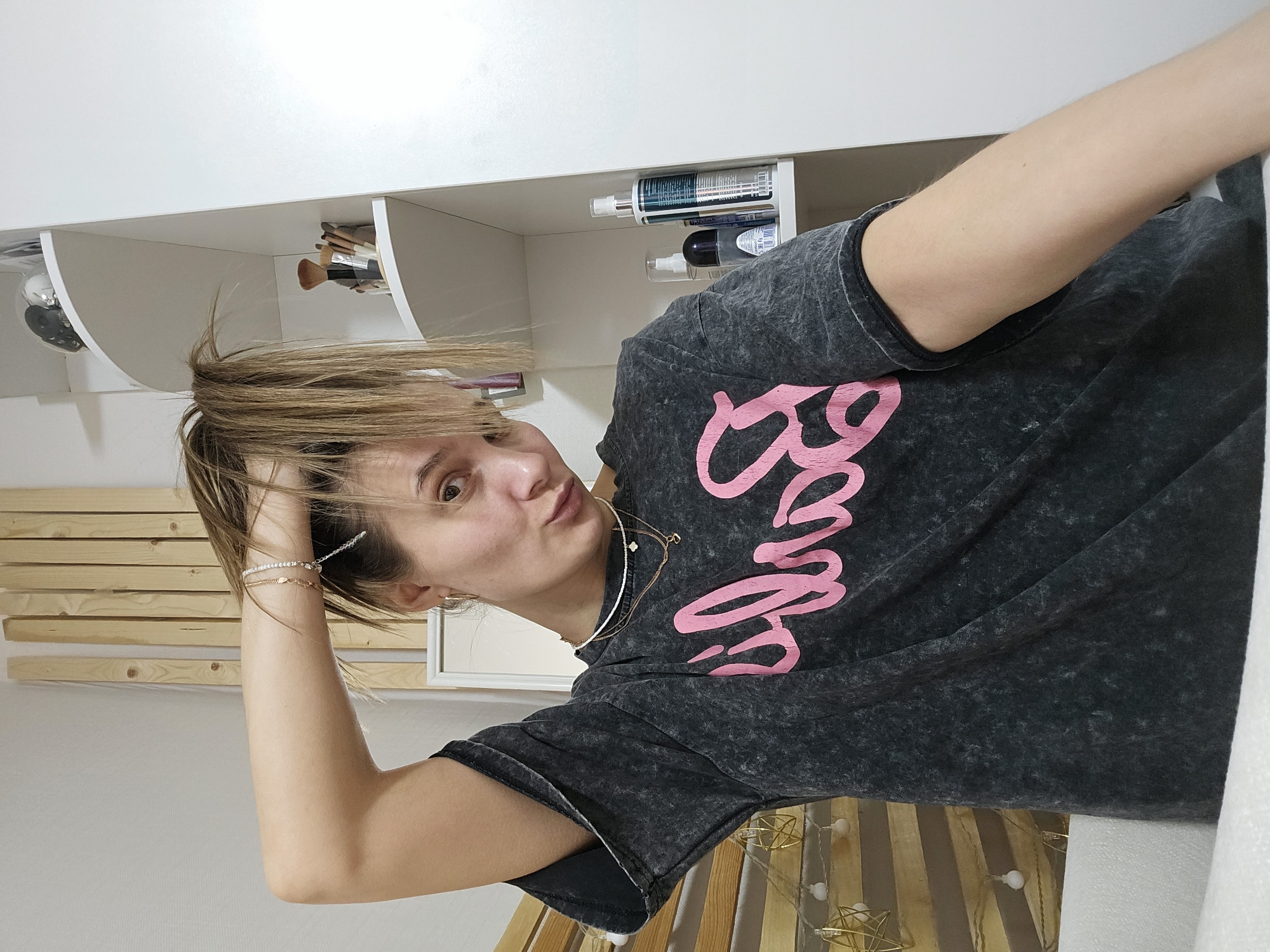 A woman is posing for a selfie in an indoor setting, possibly a bedroom. She has medium-length blonde hair and is wearing a gray t-shirt with the word "Barbie" in pink lettering. Her left arm is raised, with her hand touching her hair. She's wearing bracelets on her left wrist and necklaces around her neck.
Behind her are white shelves with various items, including cosmetic brushes, bottles, and other small objects. A wooden headboard or decorative element is visible in the background, with string lights adorning it. The location is not identifiable as Chaikovsky, Russia, based on the visual information provided.