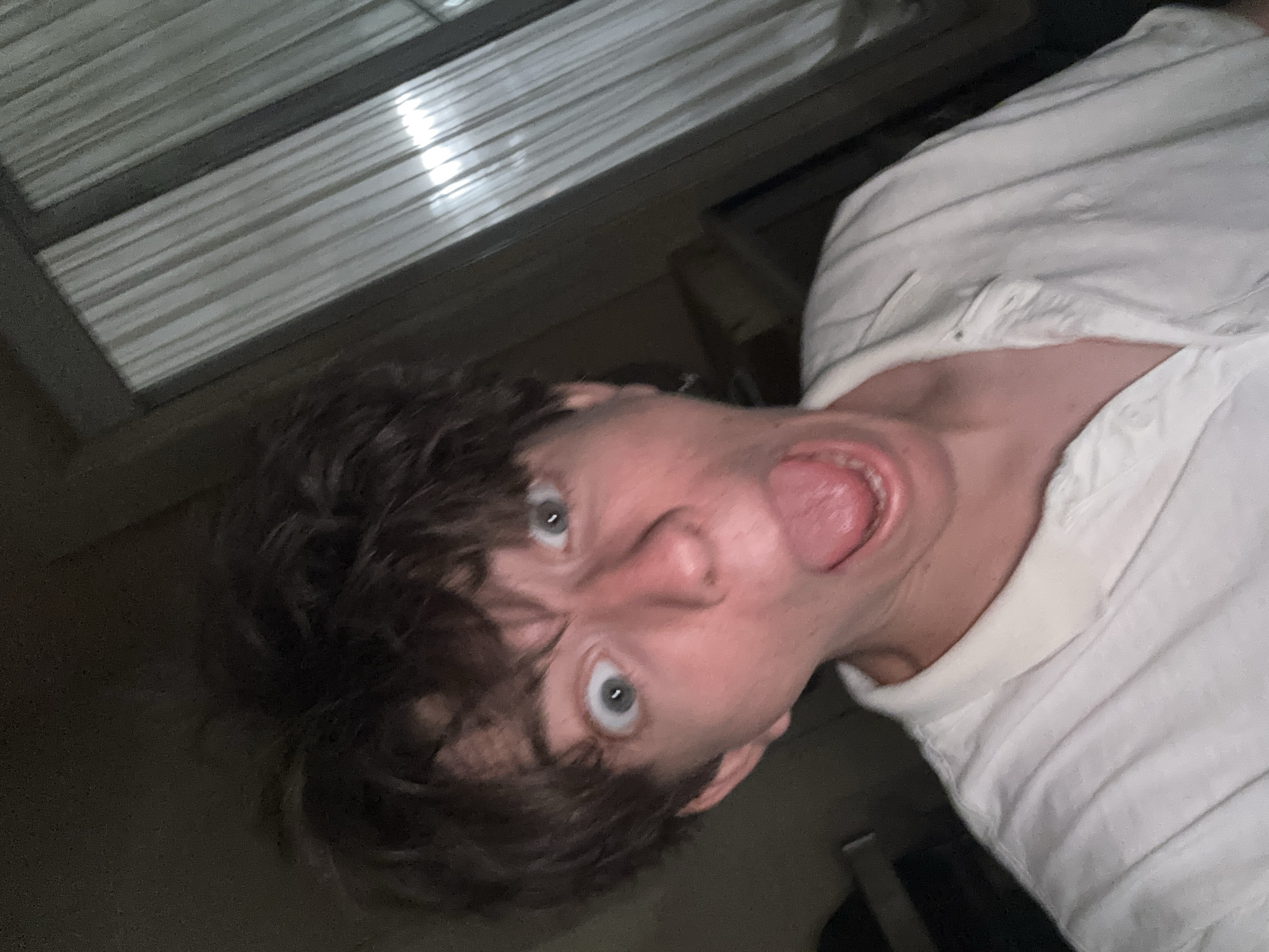 The image is a close-up, low-angle selfie of a young person with disheveled brown hair and wide, surprised eyes. Their mouth is open, with their tongue sticking out. They are wearing a white collared shirt with visible stitching. The background is dark, with a window covered by sheer white curtains on the right side, through which some light is visible. The overall impression is of a candid, playful moment. The image was taken in the city of Cupra Marittima, Italy.