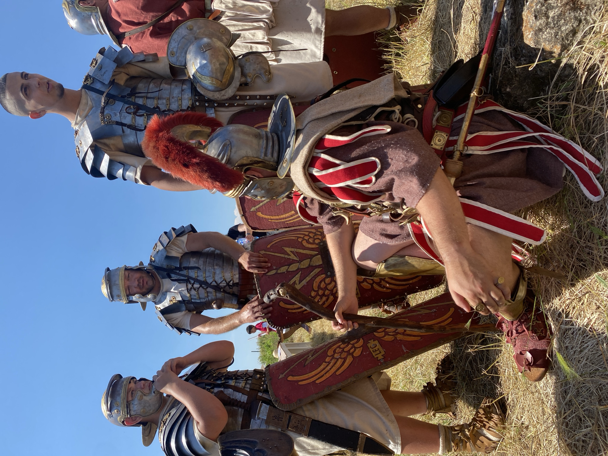 The image displays five individuals in Roman military re-enactment attire, situated outdoors on dry, golden-brown grass under a clear blue sky, identified as Gigen, Bulgaria.
In the foreground, a male figure is seated, angled with his back partially towards the viewer. He wears a dark brown tunic with distinct red and white pteruges on the shoulders and waist, and red caligae on his feet. A silver helmet adorned with a prominent red horsehair crest covers his head. He holds a wooden staff, tipped with metal, in both hands, and wears two gold-colored rings on his right hand.
To the left, a male figure stands, facing slightly right. He is equipped with a silver lorica segmentata over a light-colored tunic and a silver helmet, appearing to adjust a component of his headgear. A red scutum (shield), decorated with golden winged motifs and the inscription "LEG", rests against his left leg. A gladius scabbard is visible on his left hip, and he wears light brown caligae.
In the mid-ground, a third male figure stands facing forward, similarly dressed in lorica segmentata, a light tunic, and a silver helmet. His hands are placed on the upper edge of another red scutum.
Behind him and to the right, a fourth male figure stands, wearing lorica segmentata over a light tunic. He holds a silver helmet in his left hand, his head uncovered revealing a modern short-cropped haircut.
To the far right, a fifth male figure is partially visible, dressed in a red tunic.
The scene is brightly lit by direct sunlight, creating strong shadows. A small child in a red shirt is discernible in the distant background.