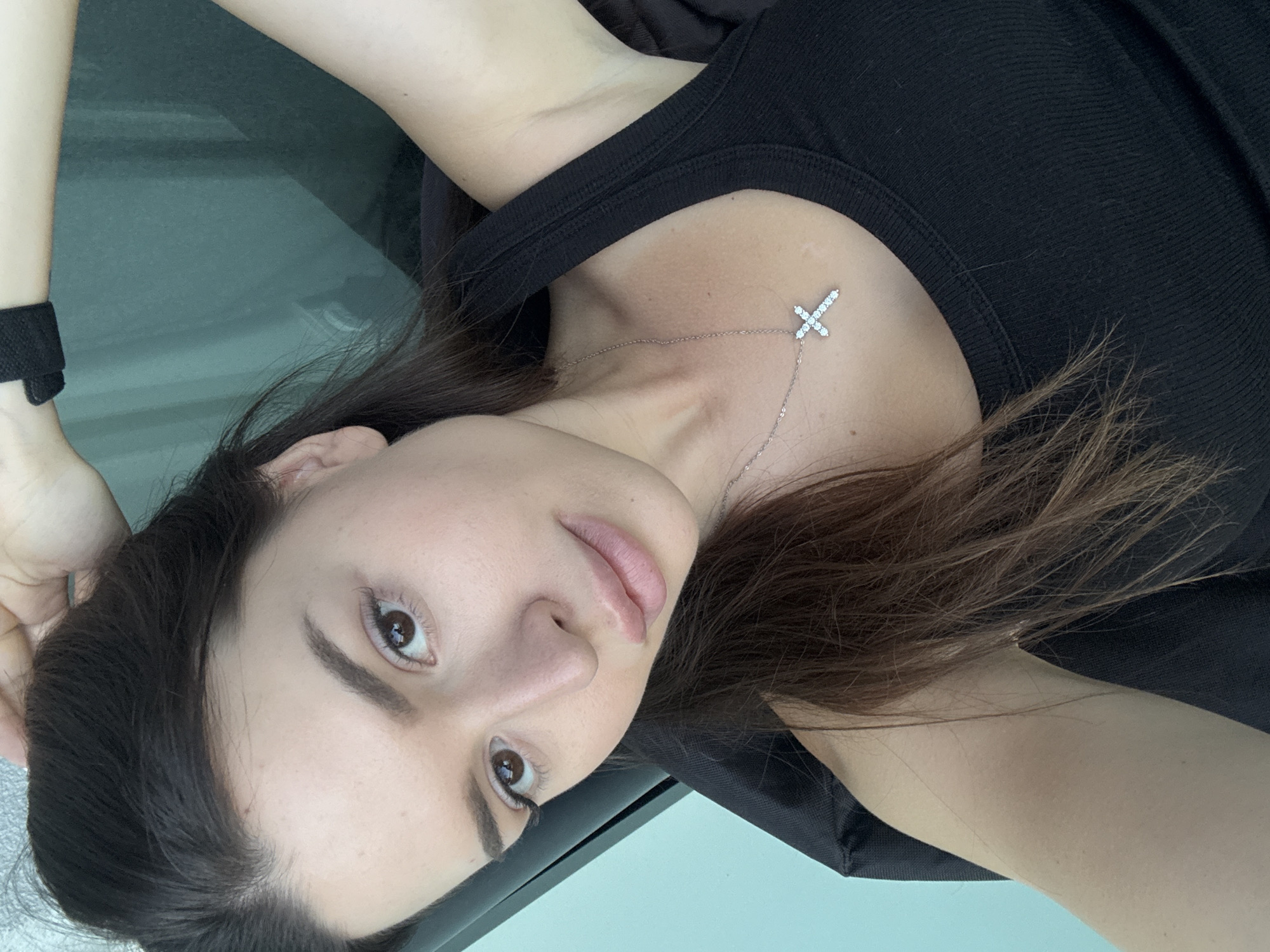 A young woman with long, dark brown hair is looking at the camera, her right hand in her hair. She is wearing a black tank top and a silver necklace with a diamond-encrusted cross pendant. The background is out of focus, but appears to be a window with a teal curtain behind it, suggesting an indoor setting. The lighting is soft and natural, casting subtle shadows on her face. The woman's pose and expression are relaxed and casual, hinting at a candid moment. The image does not provide any specific information about her location in Prague, Czechia.