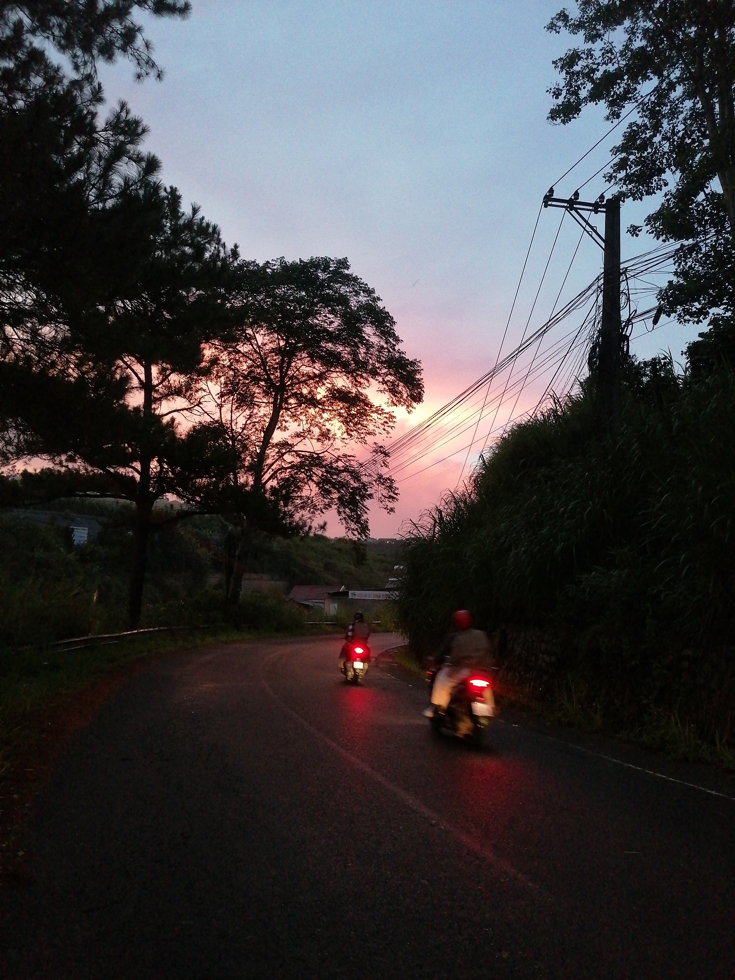 The image shows a road scene in Da Lat, Vietnam, at dusk or dawn. The sky is a mix of blue and pink hues. Two motorcycles are traveling on a winding asphalt road. The first motorcycle is further ahead, and the second is closer to the camera. Both motorcycles have their tail lights illuminated. The road is bordered by dense vegetation and trees. On the right side of the road, there is a utility pole with numerous wires. In the background, there are glimpses of buildings and hills. The overall lighting is dim, emphasizing the glow of the sky and the motorcycle tail lights.