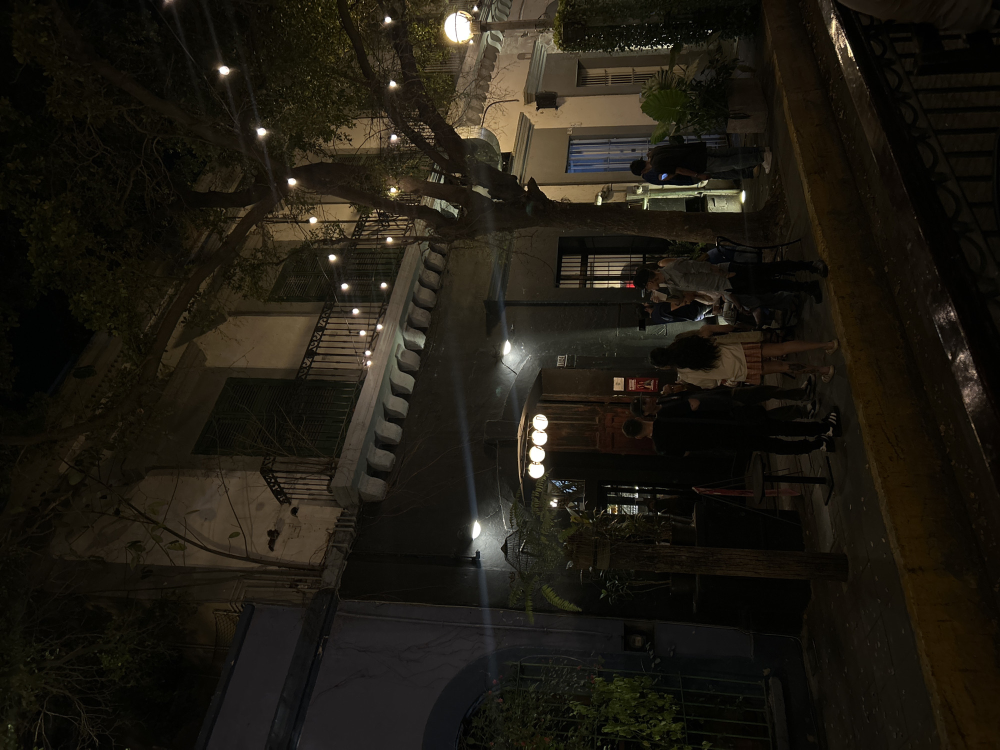 The image shows a nighttime scene in Mazatlán, Mexico. The street is dimly lit by overhead string lights and building-mounted fixtures. A multi-story building with a balcony and decorative facade is visible on the left. A tree with bare branches partially obscures the view of the building. On the right, a group of people is gathered outside what appears to be a bar or restaurant. Some individuals are standing, seemingly waiting to enter. The building is illuminated by several lights above the entrance. The sidewalk is narrow and lined with a curb. The overall ambiance suggests a lively nightlife scene.