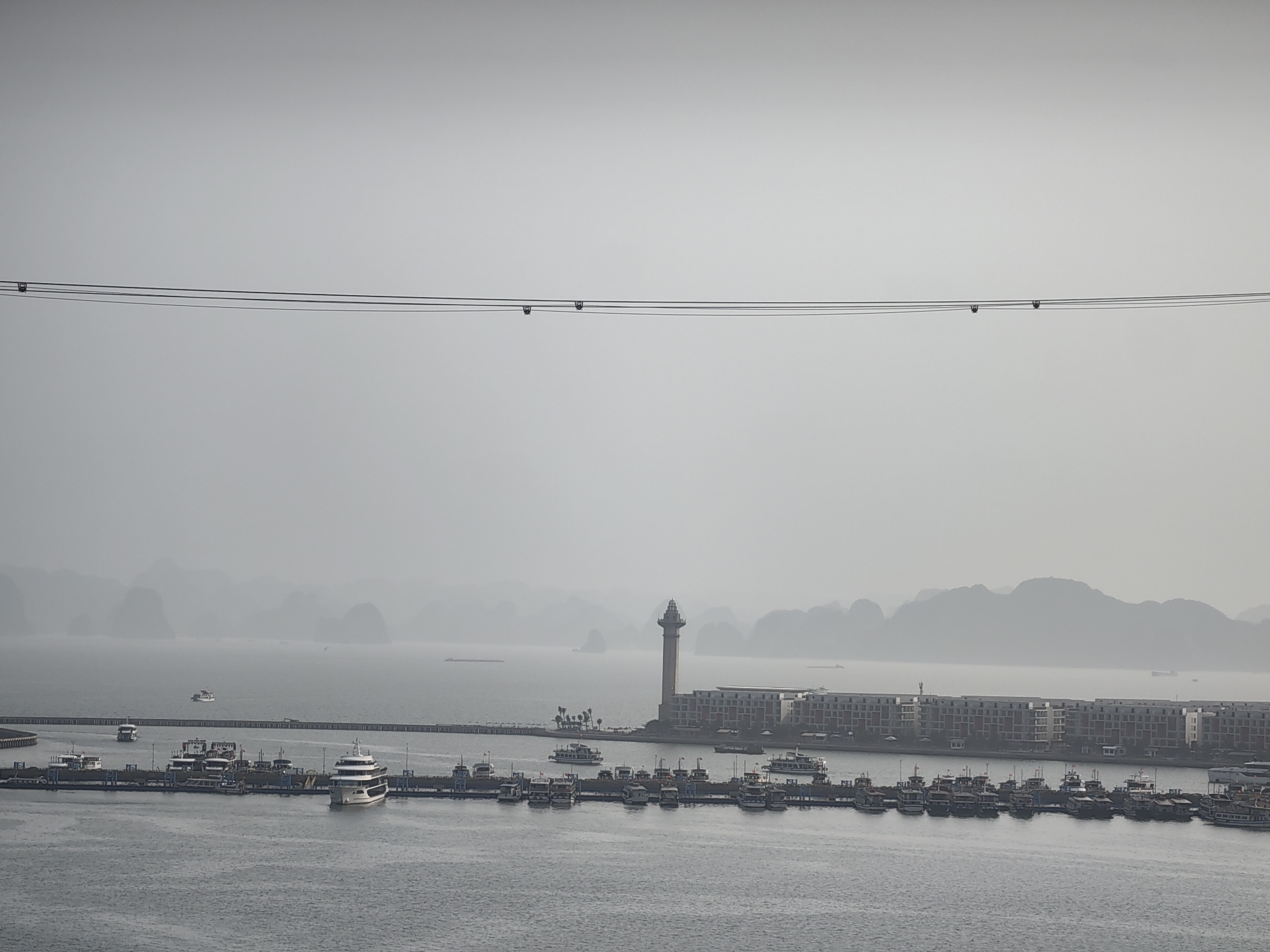 An overcast, aerial shot depicts a harbor scene in Bai Chay Ward, Vietnam. In the foreground, water fills the frame, with a large pier extending across the middle. Numerous boats are docked along the pier. Adjacent to the pier, there is a lighthouse next to a large building, and a series of rocky islands are visible in the background, fading into the mist. A pair of wires stretch horizontally across the upper portion of the image. The scene is gray and subdued, likely due to weather conditions.