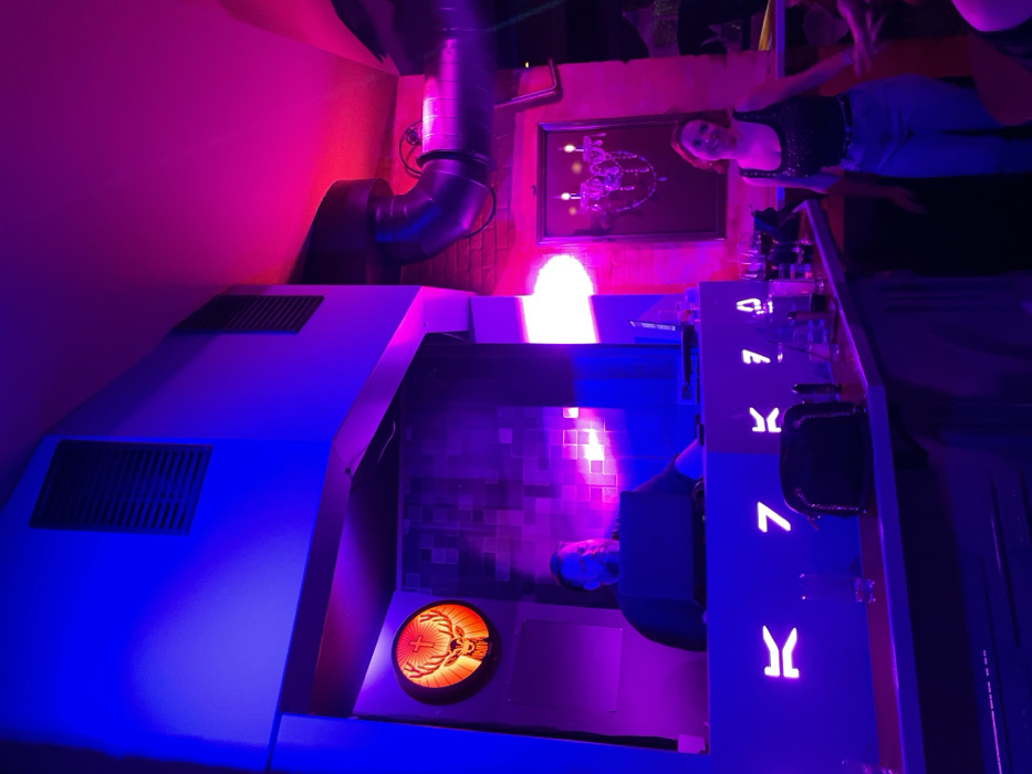 This media file captures an indoor bar or nightclub scene in Drapetsona, Greece, at night, characterized by vibrant and dynamic artificial lighting. The image is primarily bathed in strong blue, purple, and magenta/pink hues, creating a lively and modern atmosphere.
In the upper right, a woman with a smiling expression is visible, standing near a bar counter. She is wearing a dark top and light-colored pants. Adjacent to her, on the wall, hangs a framed piece of artwork or decoration that resembles an elaborate chandelier or abstract glowing object. In the lower center/right, partially obscured by the bar, another person is seated.
The prominent objects in the foreground include a large, angular structure, possibly part of the bar or a DJ booth, illuminated in blue light, featuring grilles on its surface. Further back, a bar counter stretches across the scene, adorned with several glowing, stylized 'Y'-shaped symbols or letters. On this counter, bottles and glasses are arranged. A distinct, illuminated circular sign displaying the orange and red Jägermeister logo (a stag with a cross between its antlers) is prominent on a lower section of the counter.
Industrial-style silver ventilation pipes or ducts are visible along the upper left wall, adding a raw, modern touch to the decor, which also includes sections of exposed brickwork and smooth, painted walls. A bright white light source is visible behind the main bar area, contrasting with the dominant cool tones. The overall impression is one of a trendy venue where people gather to socialize and enjoy drinks.