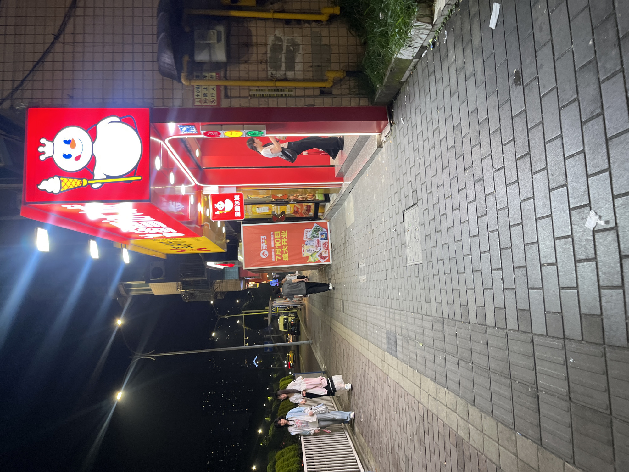 The image depicts a street scene at night in Shapingba District, China. A brightly lit store with a red facade and a prominent sign featuring a snowman character holding an ice cream cone is the central focus. The store appears to be a beverage or dessert shop.
To the left of the store, two young women are walking on a paved sidewalk. The woman on the left is wearing a light-colored jacket over a white top and blue pants. The woman on the right has a long, flowing skirt and a jacket. They are both carrying bags.
Further down the sidewalk, a person wearing a grey t-shirt and dark pants is walking away from the store. The sidewalk is constructed of rectangular pavers with a tactile paving strip for the visually impaired.
In front of the store, a woman in a white t-shirt and black pants is standing near the entrance, looking at her phone. The store entrance is recessed and illuminated by red lighting. Above the entrance, a smaller sign with the same snowman character is visible.
To the right of the store, a wall with yellow pipes and some signage is visible. The environment suggests an urban setting with streetlights illuminating the scene and distant city lights in the background. There is some greenery bordering the sidewalk on the far right. Trash is scattered on the sidewalk in various locations.