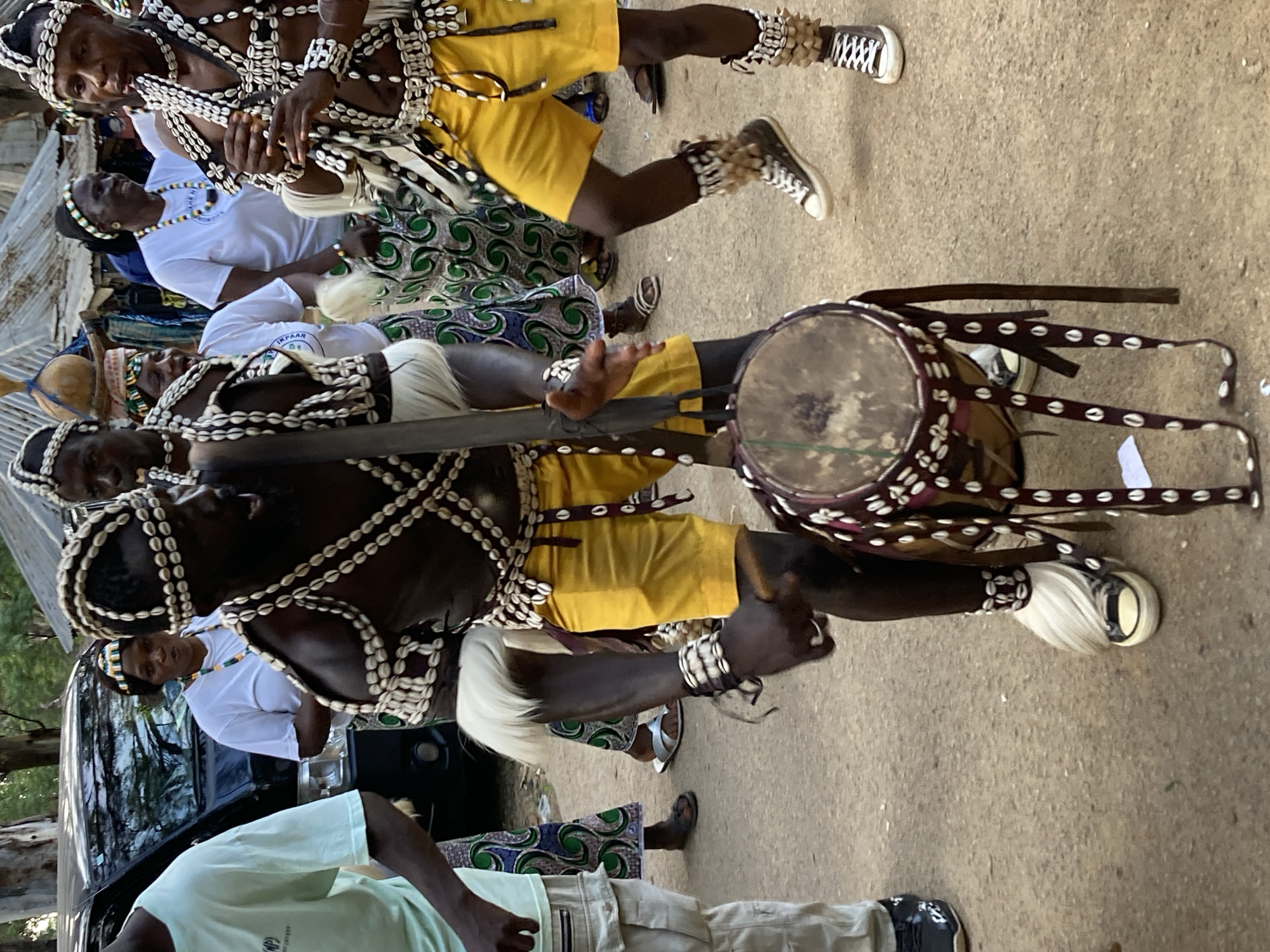 The image depicts a group of people engaged in a cultural performance, likely a procession or ceremony, in Jos, Nigeria. The central figures are adorned in traditional attire characterized by intricate shell necklaces, bracelets, and headbands. They wear yellow shorts or skirts, and some have decorative white fur attached to their legs. One individual is playing a drum, a circular object with a decorative band around its rim and a wooden handle. The ground appears to be dirt or gravel, suggesting an outdoor setting. In the background, a car is partially visible, along with hints of trees and structures, possibly a market or a village. The lighting suggests it is daytime, and the atmosphere appears festive or ceremonial. No visible text is present in the image.
