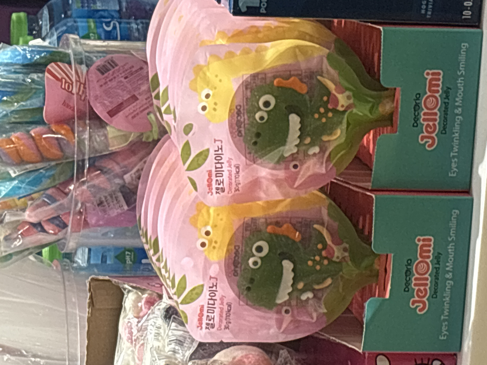 The image depicts a retail display of individually packaged jelly candies, specifically JellOmi brand "Decorated Jelly" featuring dinosaur designs. The candies are presented in pink packaging with green and yellow dinosaur characters, and the packaging is housed in teal cardboard boxes. The background shows a variety of other confectionery items, including lollipops and bottled beverages, suggesting a candy store or supermarket aisle. The lighting is bright and the overall scene is well-lit, typical of a retail environment. There are no people visible in the image. The specific location of this display within Dubai, United Arab Emirates, cannot be determined from the provided image alone.