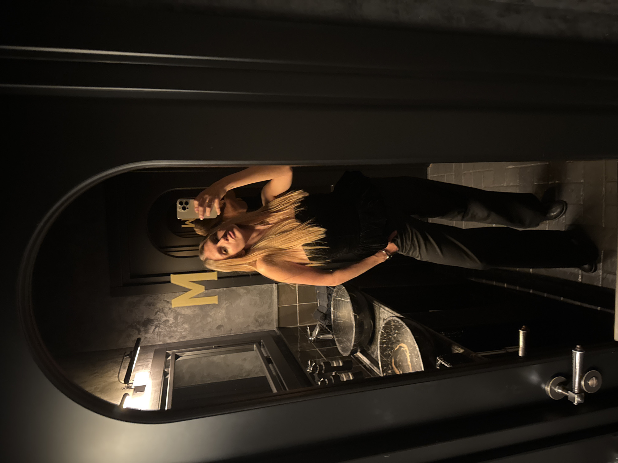 A blonde woman is taking a selfie in a mirror in a dimly lit bathroom. She is wearing a black sleeveless top and black pants. The bathroom has two sinks with a marble pattern. The mirror is framed in black. In the background, a gold letter "M" is visible on the wall. The location is likely in Tbilisi, Georgia, based on other image data.
