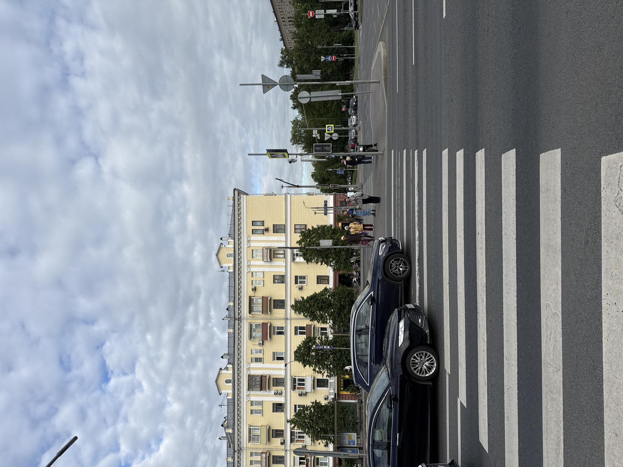 This image depicts an urban street scene in Moscow, Russia, centered around a pedestrian crossing at an intersection. The perspective is taken from slightly above, looking across the street.
On the left side of the road, a tall, multi-story building with a light yellow facade stands prominently. It features numerous windows, some with small balconies, and decorative architectural elements. On its ground floor, a shop sign displaying the Cyrillic word "ЧАЙ" (Chai, meaning Tea) is visible. Trees with green foliage line the street in front of the building.
The road itself is asphalt, featuring a wide pedestrian crosswalk with distinct white "zebra" stripes. Several dark-colored cars are visible, stopped or parked along the right side of the road next to the crosswalk. Numerous traffic poles stand at the intersection, adorned with various road signs (including yield and no-entry signs), traffic lights, and pedestrian signals.
Several pedestrians are present in the scene. Some individuals or small groups are standing on the sidewalk near the building, while others are on or approaching the crosswalk, indicating an everyday activity of crossing the street. They are dressed in typical casual clothing.
The sky is heavily overcast with white and gray clouds, suggesting a cloudy day without direct sunlight. The overall atmosphere is calm and mundane, characteristic of a routine daytime street environment. No specific time of day is indicated beyond daylight, likely mid-morning or afternoon given the diffused light.
