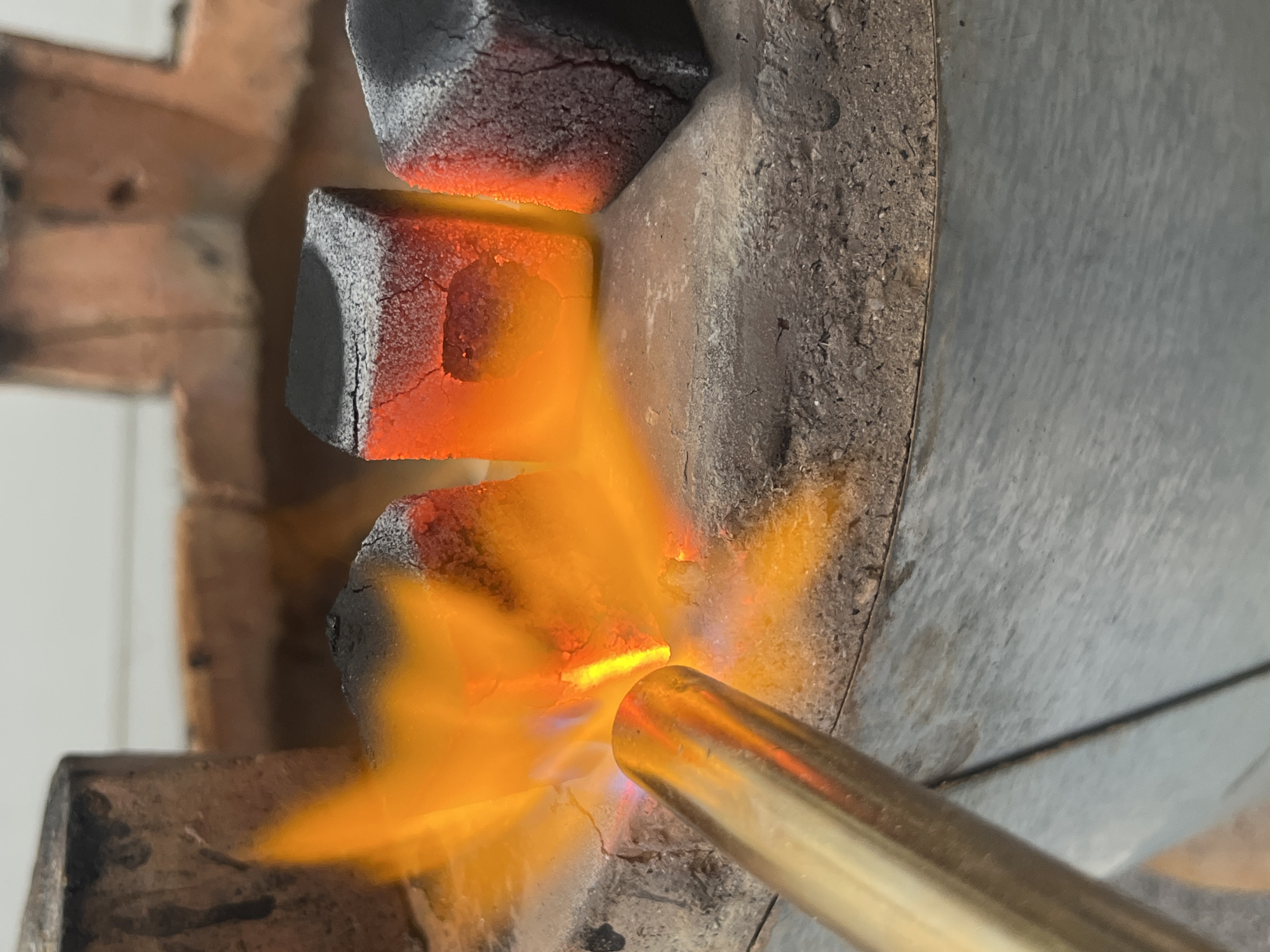 A close-up shot shows a butane torch igniting cube-shaped coals in what appears to be a metal container or furnace. The flame is a vibrant yellow-orange, engulfing the bottom coal and licking up to the middle one. The coals are textured and cracked on the surface. The metal container has a rough, speckled texture. The setting could possibly be in Bac Nha Trang Ward, Vietnam.