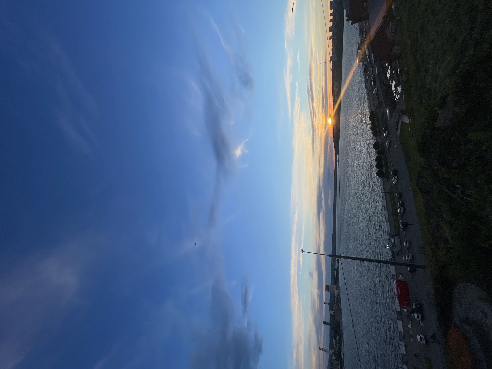 The image depicts a sunset over a body of water, likely a river or bay, bordered by an urban landscape. The sun is positioned on the horizon, emitting a bright glare that reflects on the water's surface. Buildings and structures are visible along the shoreline, suggesting an industrial or commercial area. A road runs parallel to the water, populated with parked cars and possibly some vehicular traffic. Vegetation, possibly grass or trees, is present along the embankment. The sky is a gradient of blues and oranges, indicative of the sunset. The location is a city of unknown name and country.