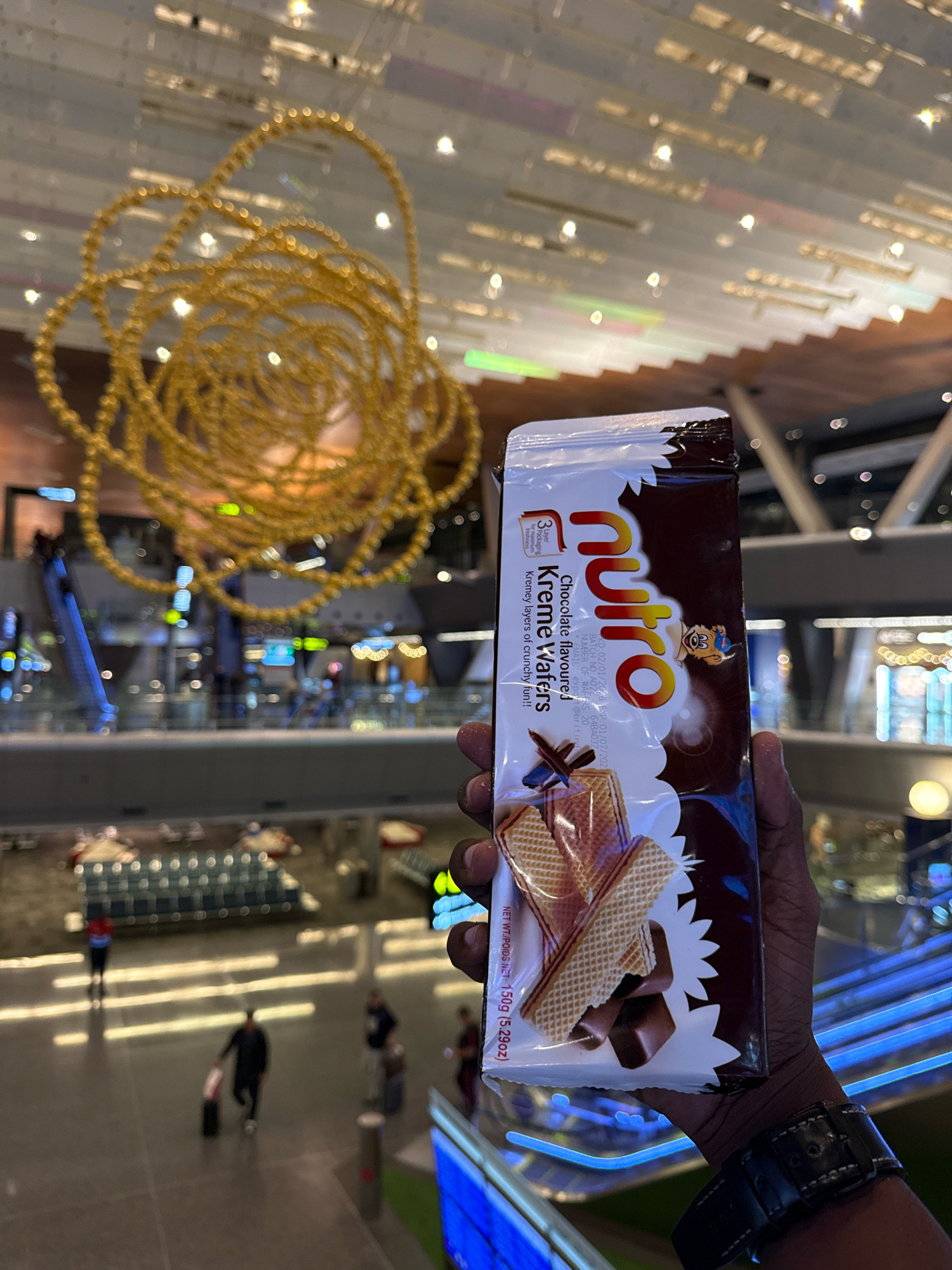 In Doha, Qatar, a person holds a package of Nutro Kreme Wafers. The package is predominantly white with brown lettering and features an image of wafers and chocolate. Behind the person, a large, golden, abstract art installation hangs from the ceiling. The background reveals the interior of a modern airport terminal with escalators, seating areas, and several people walking. The lighting suggests an indoor environment.