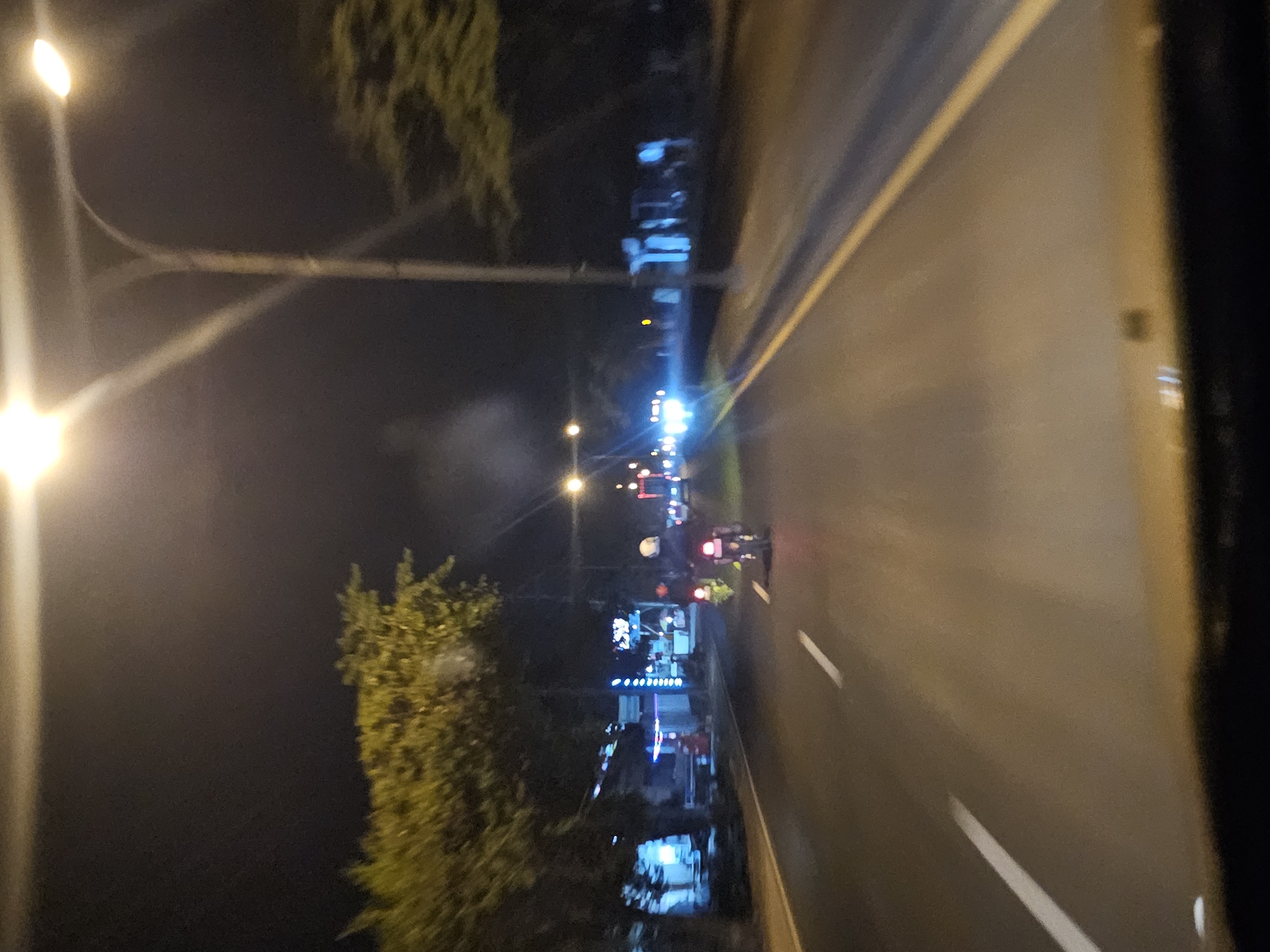 The image captures a nighttime scene on a road in Waruwetan, Indonesia. Two individuals on motorcycles are visible, one slightly ahead of the other. They are illuminated by streetlights and the headlights of passing vehicles. The road is paved and marked with white lines. On the left side of the road, there are trees and buildings with illuminated signs, suggesting a commercial or residential area. The overall ambiance is one of a typical urban street at night, with the distinct glow of artificial lights against the dark sky.