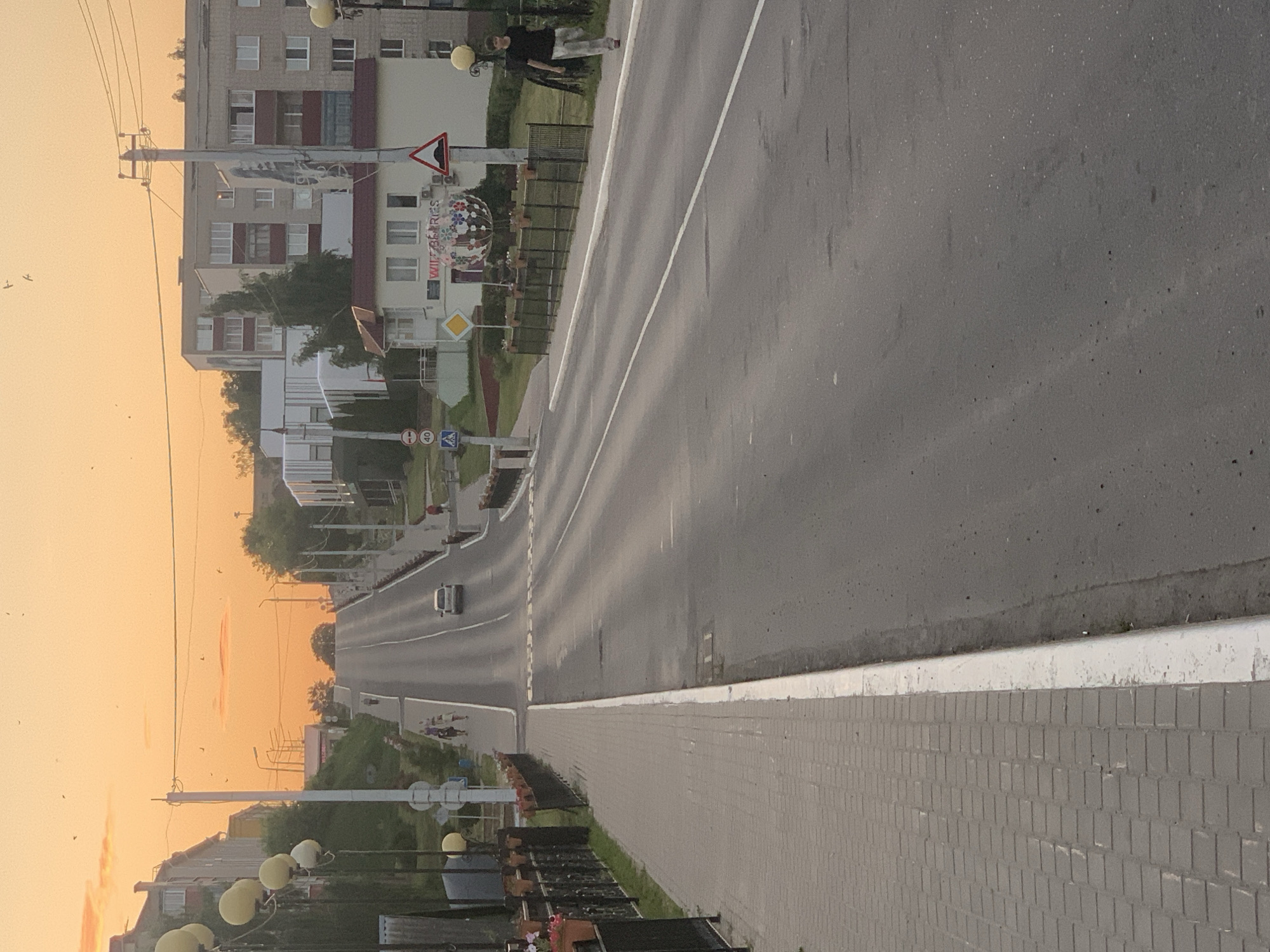 The image depicts a street scene in Kapyĺ, Belarus, during what appears to be sunset or sunrise, given the warm orange hues of the sky. The photograph is oriented vertically, with the road and sidewalk dominating the right side, and buildings and the sky on the left.
In the foreground, a sidewalk paved with light gray bricks runs along the edge of a dark asphalt road. A white curb separates the road from the sidewalk. Streetlights with round yellow globes are visible along the sidewalk.
Further down the road, a white car is driving away from the camera. The road has distinct white lane markings. On the left side of the road, there are several multi-story buildings, typical of an urban environment. One building has a facade with visible windows and balconies. A decorative spherical object with a mosaic-like pattern is positioned in front of one of the buildings. Traffic signs are also present, including a red and white triangular sign with a black symbol, and a yellow diamond-shaped sign.
In the sky, there are a few birds flying. The overall atmosphere suggests a peaceful evening or morning in a residential or commercial area. There is no discernible emotional tone, and the image focuses on capturing the scene itself. There is some text visible on the decorative sphere, which appears to read "WILD BERRIES". There are also some numbers on traffic signs, such as "40".