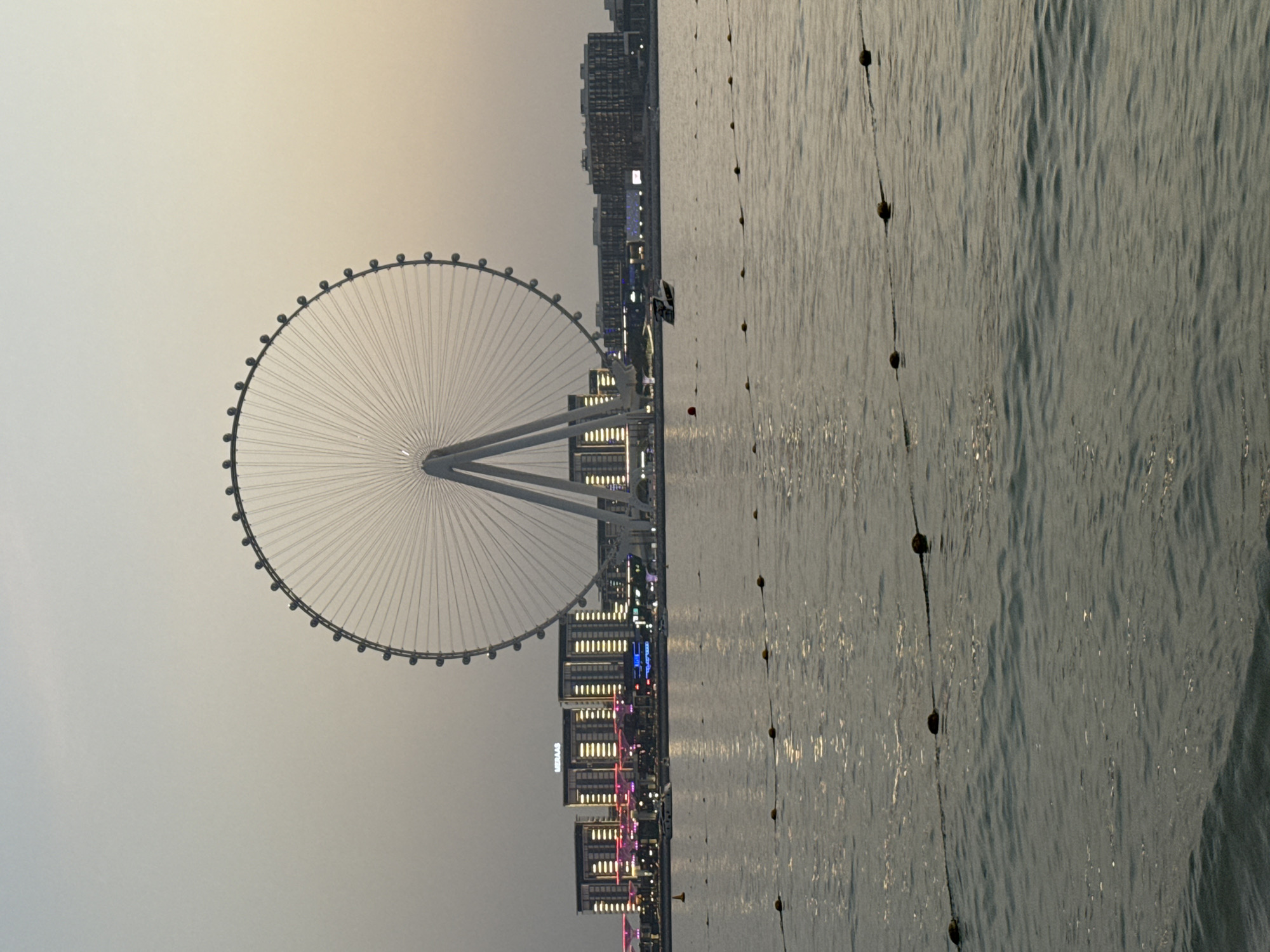 A tall Ferris wheel, the Ain Dubai, dominates the background of this image, situated on Bluewaters Island in Dubai, United Arab Emirates. The Ferris wheel is predominantly white with a wide, circular structure supported by two large A-frame legs. Numerous cabins are visible around the circumference of the wheel, many illuminated with a soft, white light.
In front of the Ferris wheel, a line of modern buildings forms the Dubai Marina skyline. These buildings are characterized by their rectangular shapes, illuminated windows, and some with colored lighting, including pink and blue accents. The name "LIERAAS" is visible on one of the buildings.
The foreground of the image consists of a body of water, likely the Arabian Gulf, reflecting the sky and the lights from the buildings. Several rows of yellow buoys are strung together by ropes, floating on the surface of the water, marking a boundary or section. A small white boat is visible in the middle distance, moving across the water. The sky above is a pale, hazy gray, suggesting either early morning or late evening light. The overall scene depicts a waterfront view with iconic Dubai landmarks.