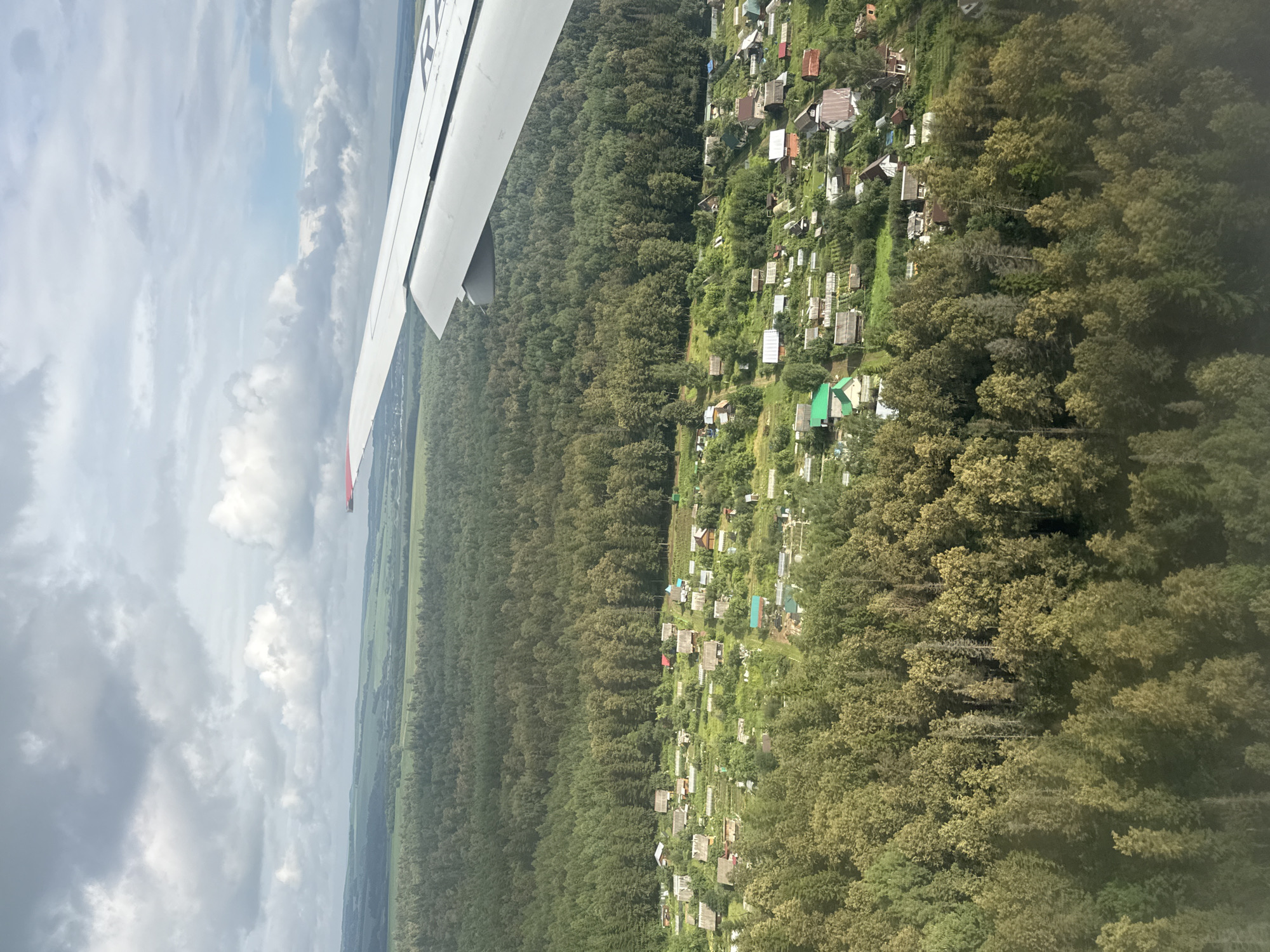 An aerial view captures a rural settlement nestled amongst dense, green forests, with an airplane wing visible in the upper right quadrant. The settlement, appearing to be in the vicinity of Vozhoi, Russia, is characterized by numerous small houses with distinct roofs, surrounded by gardens and small plots of land. The terrain slopes gently, with patches of lighter green suggesting open fields or cultivated areas interspersed with the darker, more varied shades of the forest canopy. The sky above is partially covered by dynamic clouds, with hints of blue sky visible. The image provides a snapshot of a semi-rural landscape from an elevated perspective, likely during daytime, under mixed weather conditions.