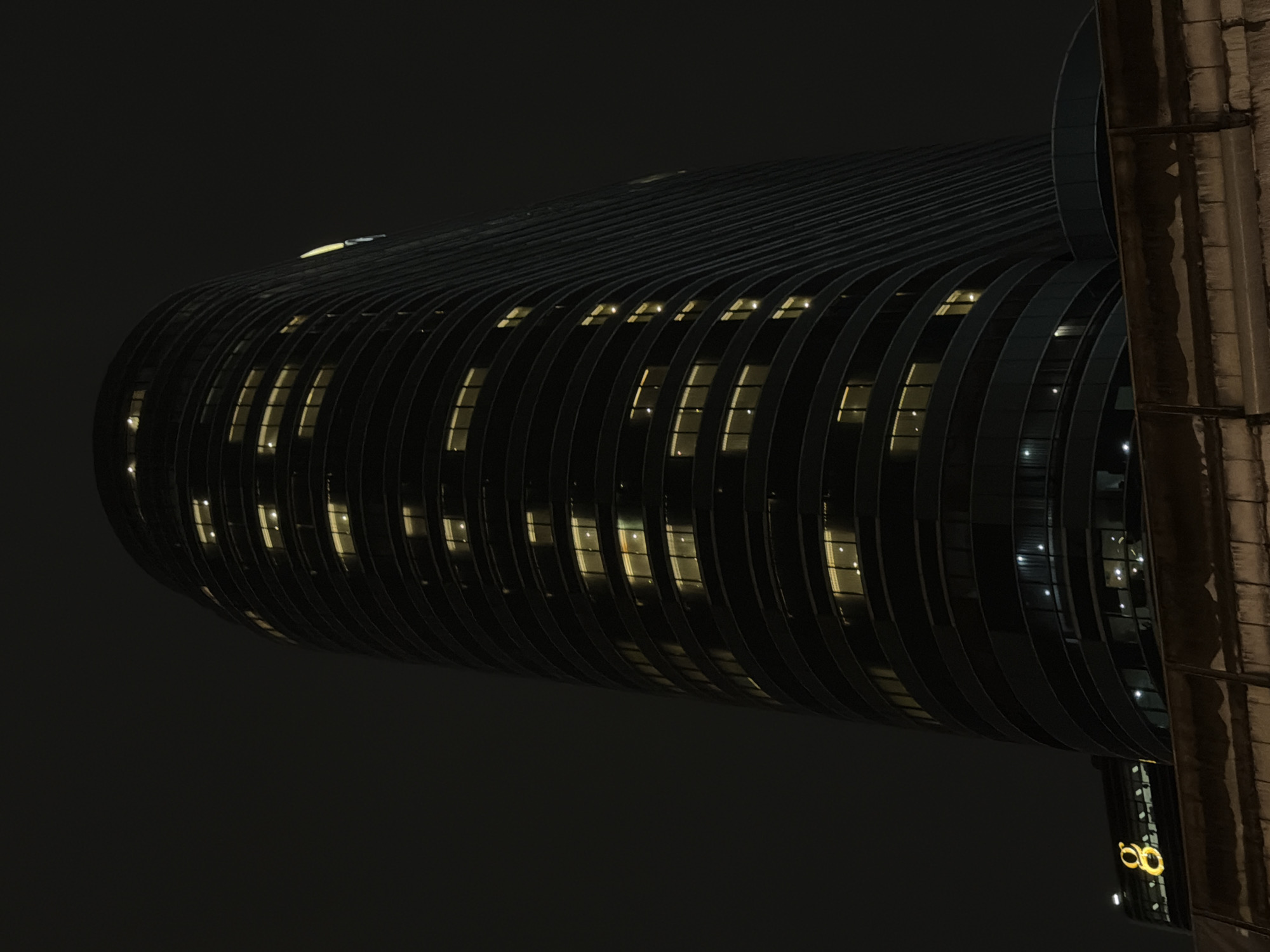 A low-angle, long shot captures a dark, cylindrical skyscraper at night in Bangkok, Thailand. The building's exterior is lined with horizontal bands of illuminated windows, creating a textured effect against the dark sky. On the lower left side of the frame, a smaller, dark building features a large, glowing golden number "8" sign. In the foreground, a rough, textured wall or barrier is visible. The overall scene is dark and moody, illuminated primarily by the artificial lights of the buildings. There are no people or visible activities depicted in the image.