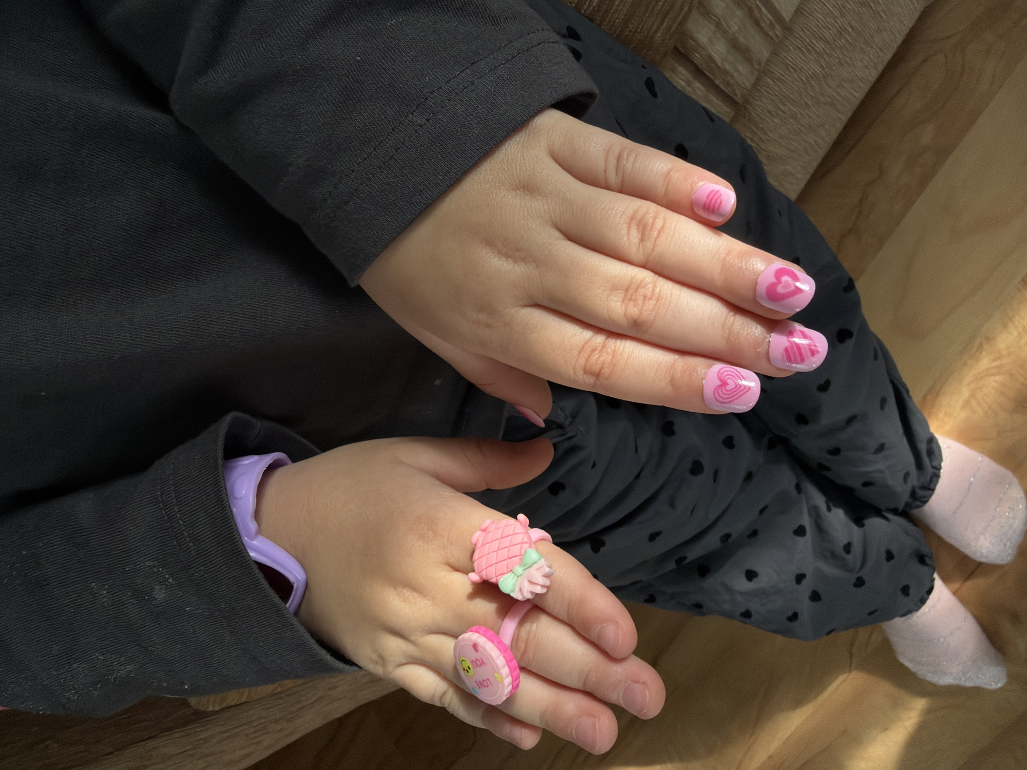 A child is seated indoors. She wears a dark gray long-sleeved shirt, dark gray pants with small black hearts, and light pink socks. Her fingernails are painted pink, some with a pink heart design. She wears two pink plastic rings on her left hand. A light purple plastic bracelet is visible under the sleeve of her shirt on her left wrist. The floor is light-colored wood.