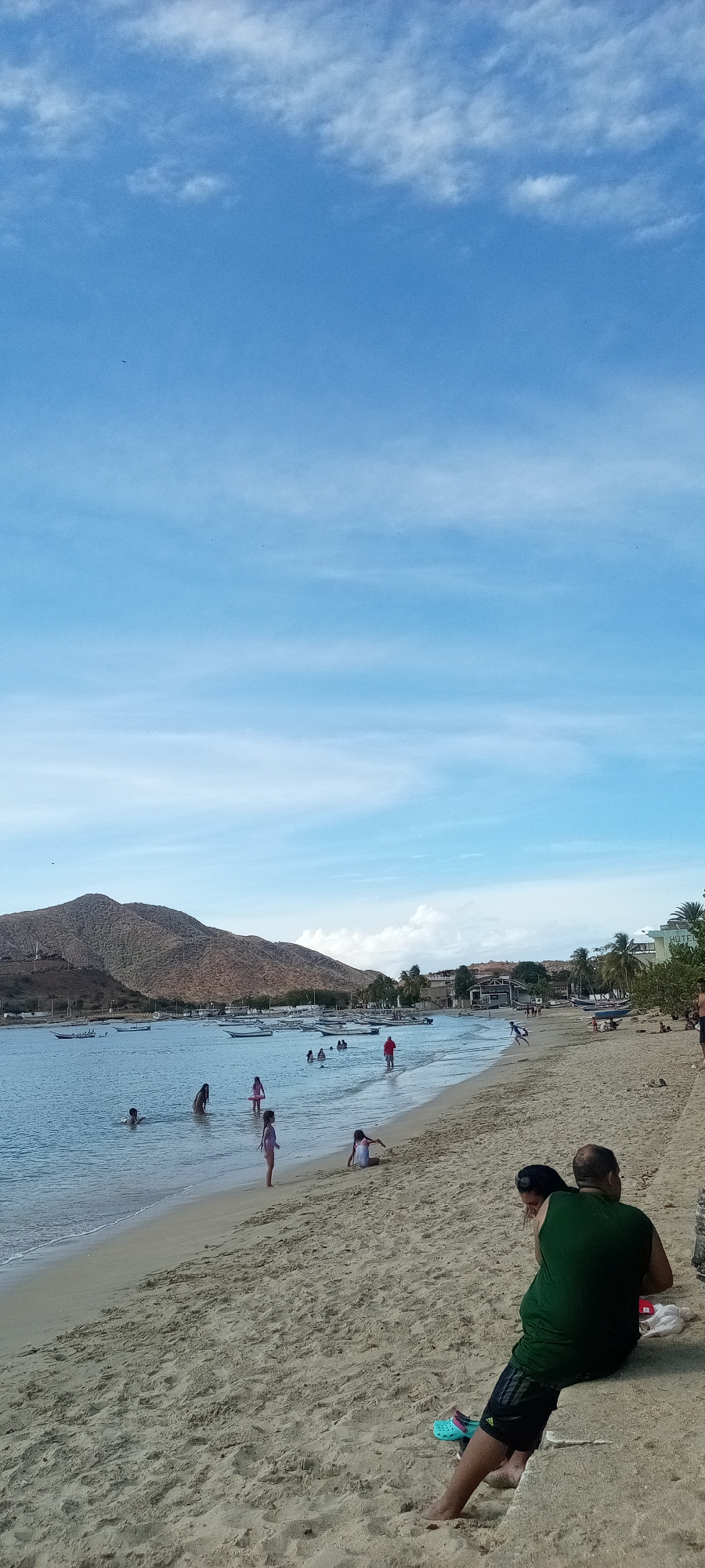 The image shows a beach scene in an unidentified location. The sky is blue with light clouds. In the background, a mountain is visible across the water. Several boats are anchored in the bay. In the foreground, people are swimming and playing in the water. Others are on the sandy beach. In the lower right corner, two people are sitting on a concrete wall, with their backs to the camera. The setting is a beach with people enjoying a sunny day.