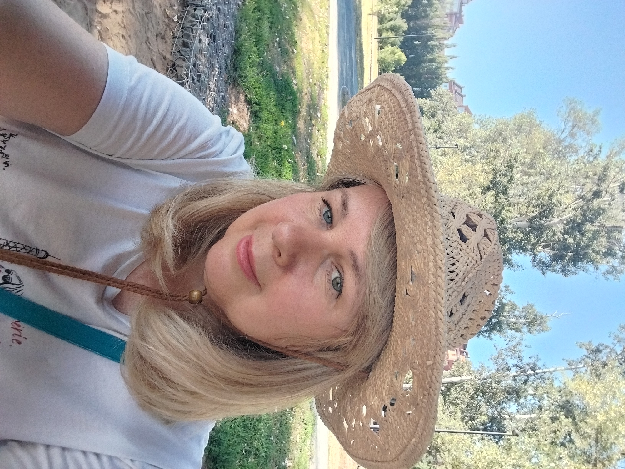 A woman wearing a straw hat and a white t-shirt is pictured in an outdoor setting. She has blonde hair and is looking towards the camera with a slight smile. The background shows trees, green grass, and a road, suggesting a park or suburban area. The image appears to be a selfie. The location is identified as Orenburg, Russia.