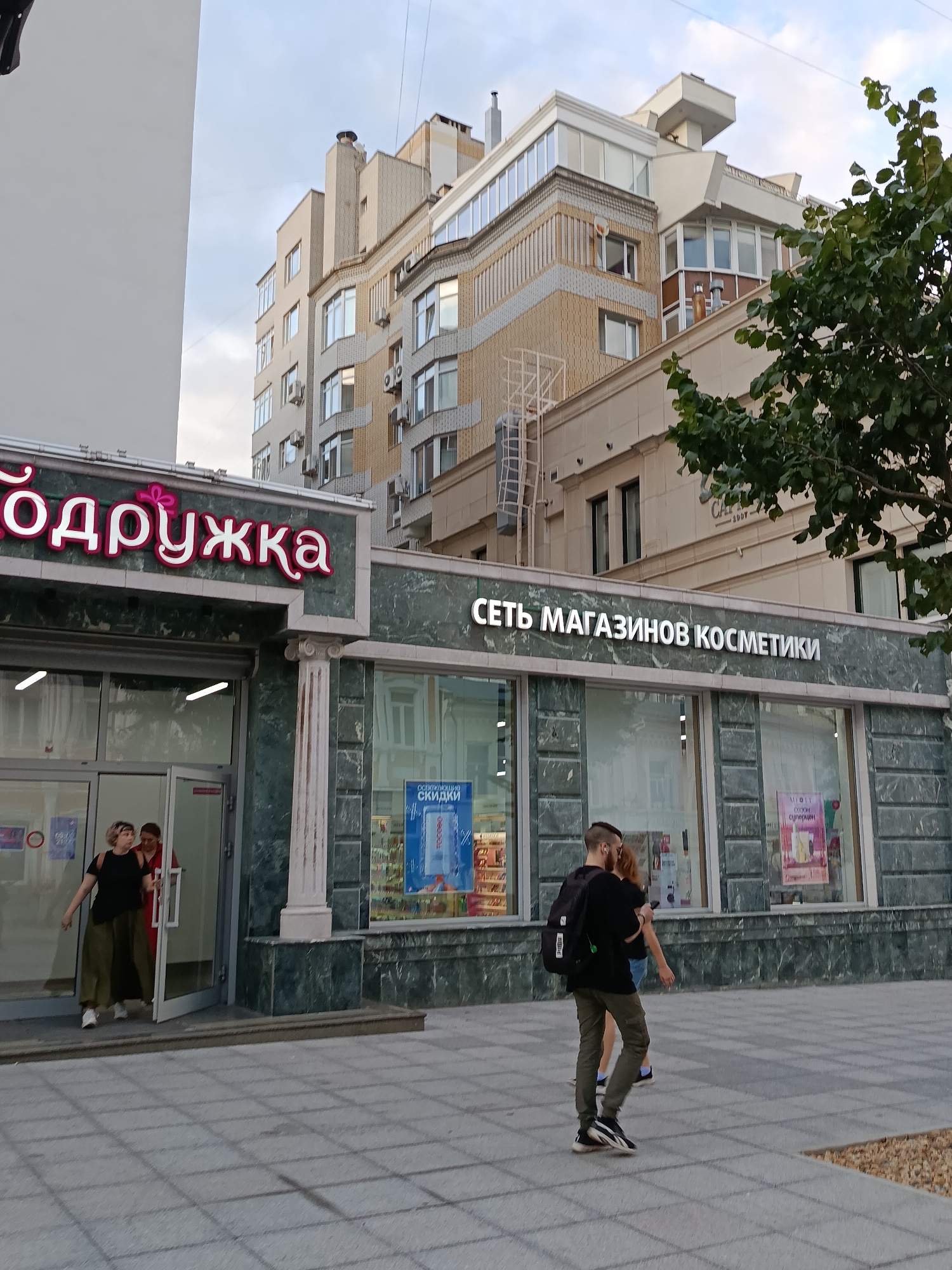 The image depicts the exterior of two adjacent stores on a city street. The store on the left has a pink neon sign with Cyrillic text reading "ПОДРУЖКА" (PODRUzhKA), which translates to "Girlfriend" or "Friend" and appears to be a gift shop. The store on the right has a similar marble facade and a white illuminated sign that reads "СЕТЬ МАГАЗИНОВ КОСМЕТИКИ" (SET' MAGAZINOV KOSMETIKI), meaning "Cosmetics Store Chain."
In front of the "PODRUzhKA" store, a woman with blonde hair wearing a black top and olive green pants exits the store, while another woman in a red outfit stands just inside the doorway. A man with a backpack, wearing a black t-shirt and dark pants, walks past the cosmetics store, with his arm around a woman who is wearing a black top and blue shorts. The street is paved with gray tiles. In the background, several multi-story buildings are visible, one of which has a distinctive architectural style with beige and grey stone cladding. A large green tree with dense foliage is on the right side of the frame, partially obscuring the upper floors of the buildings. The sky is overcast with some thin clouds. The image was captured in Saratov, Russia.