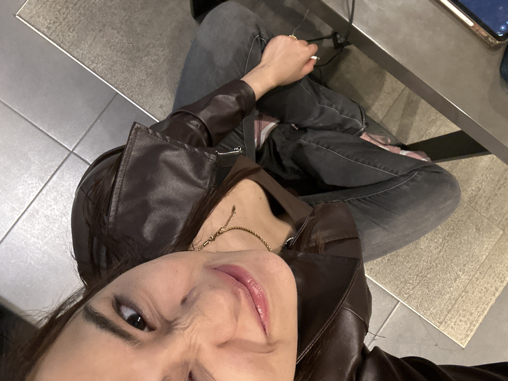 This high-angle selfie captures a person, likely a woman, seated indoors in San Andrés Cholula, Mexico. The subject is wearing a dark brown, leather-look jacket over a lighter brown top and dark grey jeans. A gold-toned chain necklace is visible around her neck. Her right hand, adorned with a gold-colored ring, holds a lit cigarette, suggesting she is smoking. She is seated with her legs crossed.
The subject's face is prominent, featuring a playful expression with a slight smirk and crinkling around her left eye, indicating amusement or a quirky pose. Her lips are slightly parted, showcasing a glossy, reddish-pink lip color. Her long, dark hair falls around her shoulders.
The background reveals an indoor environment with a dual-textured floor: light grey tiles and a section of light brown, textured mat or carpet. To the right, a dark grey table or counter surface is visible, on which a portion of a mobile phone or electronic device rests. Wires are also noticeable near the base of the table, hinting at a power source or charging station. The lighting appears to be artificial, typical of an indoor commercial or public space. There is no visible text in the image.