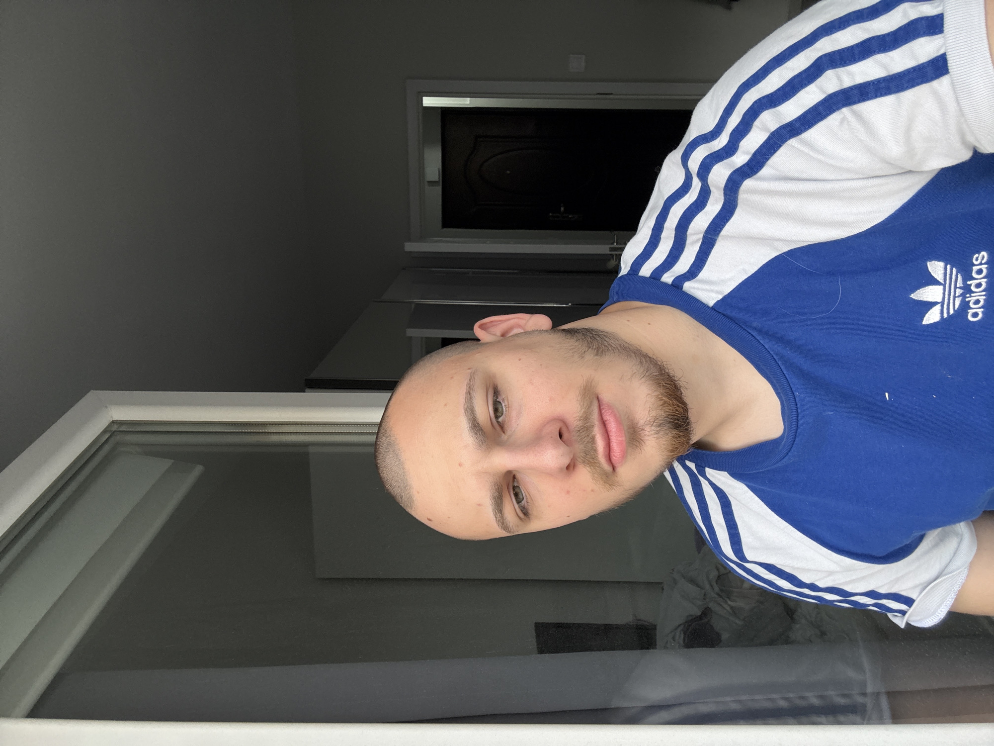 A medium shot depicts a person indoors, wearing a blue and white Adidas t-shirt. The person has short hair, light skin, and a beard. Behind them, there's a window on the left and a dark-colored door in the background. The lighting appears to be dim, suggesting it could be night or a room with limited natural light. There is no indication of the location being Stara Zagora, Bulgaria.