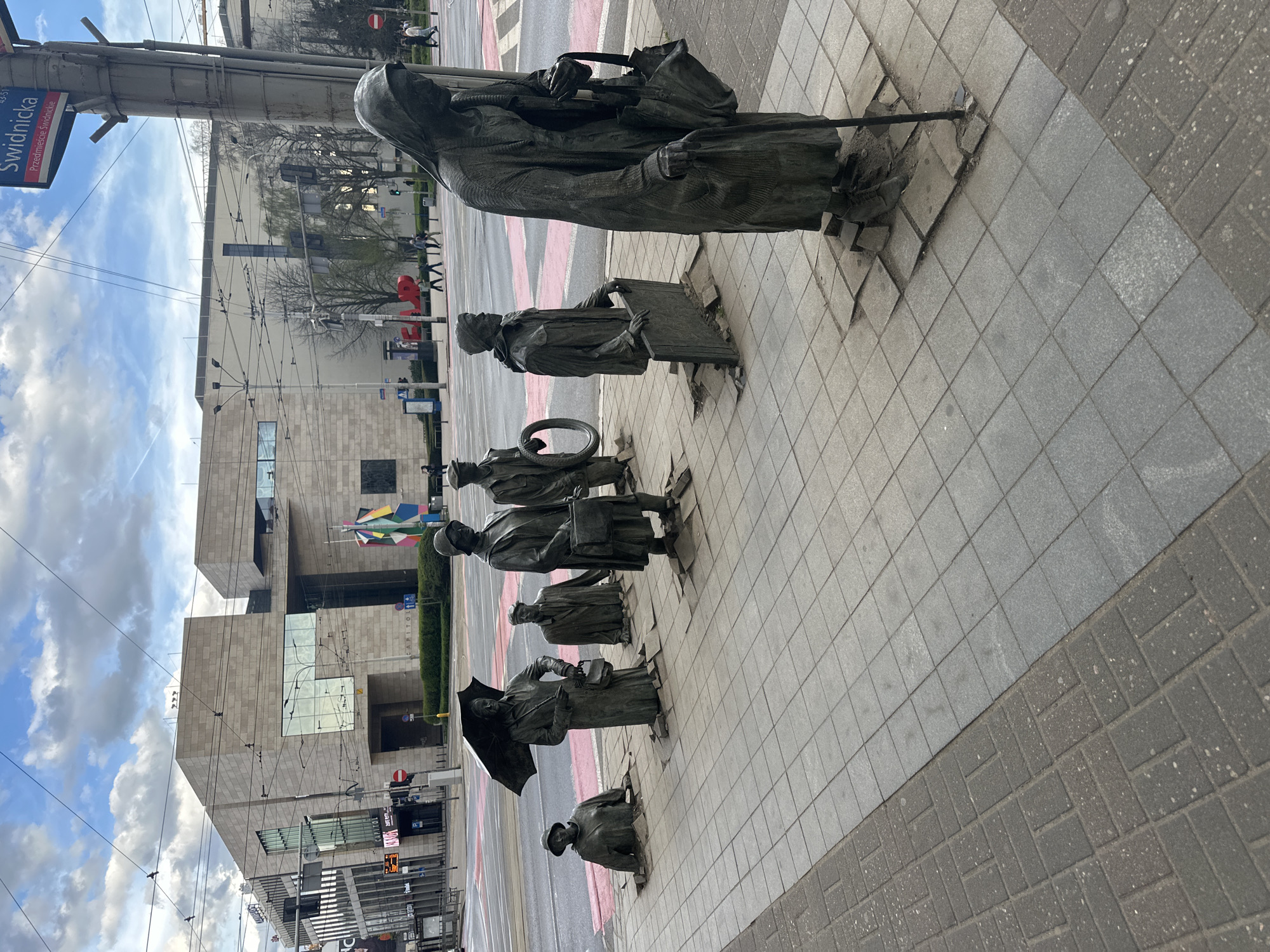 The image depicts a series of bronze statues emerging from the sidewalk on Świdnicka Street in Wrocław, Poland. The statues represent people of various ages dressed in clothing from different eras. They are situated on a grey-tiled sidewalk with a contrasting dark brick pattern along the edge. In the background is a modern building with glass and stone façade, and a street sign is visible on a pole. The sky is partly cloudy.