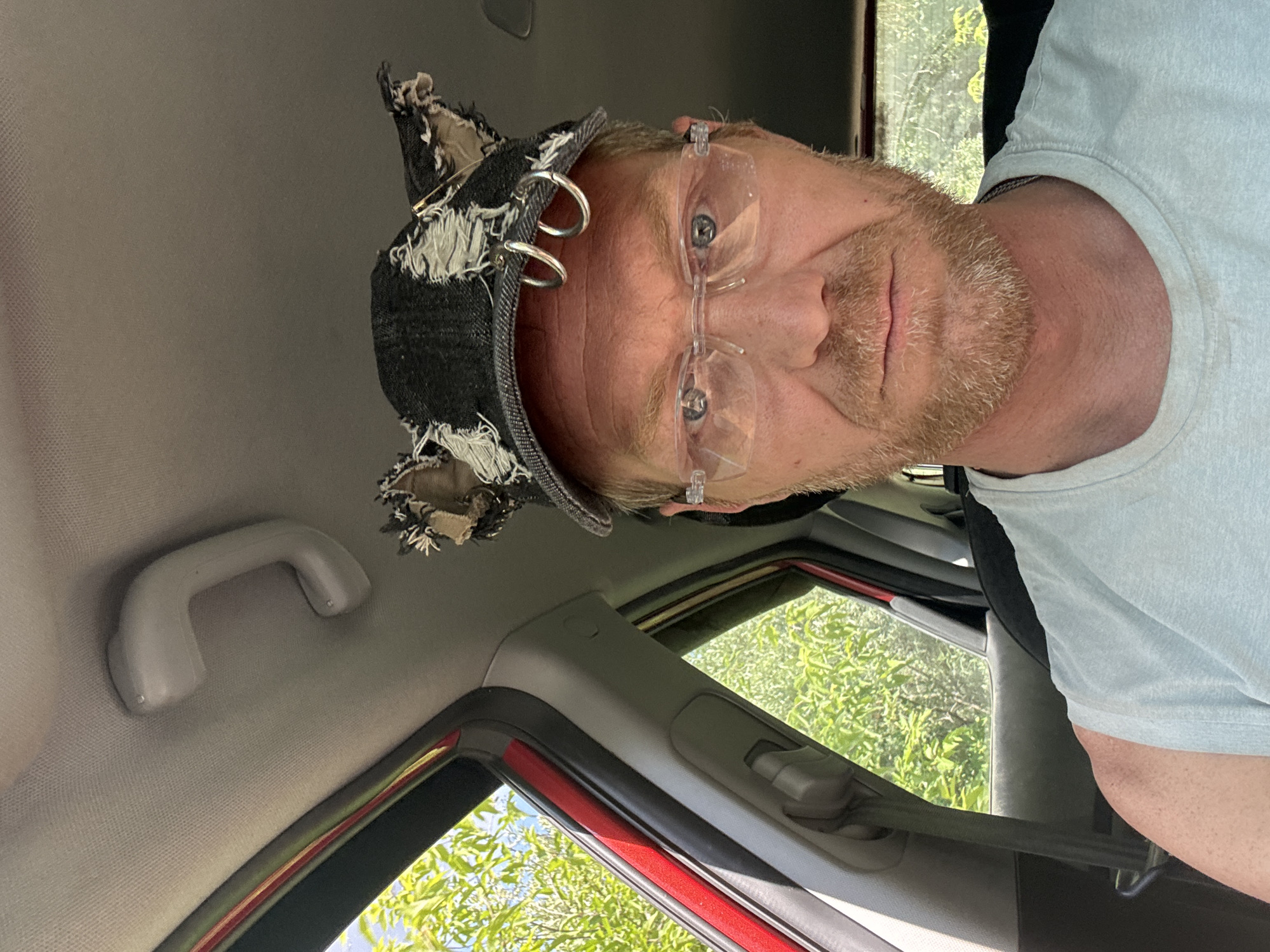 A man with a reddish-brown beard, wearing clear-rimmed glasses and a light blue t-shirt, is seated inside a vehicle. He is wearing a dark denim baseball cap that has been modified with ripped fabric and two silver hoop earrings attached to the front. Outside the vehicle, green foliage and a blue sky are visible through the windows. The interior of the car appears to be a typical passenger vehicle. No specific landmarks or indicators place the image within Chaikovsky, Russia.